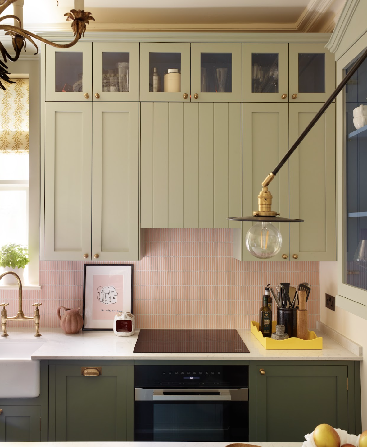 Kitchen with green cabinets, pink tile backsplash, white countertop, gold faucet, framed artwork, potted plant, and yellow tray holding bottles and utensils.