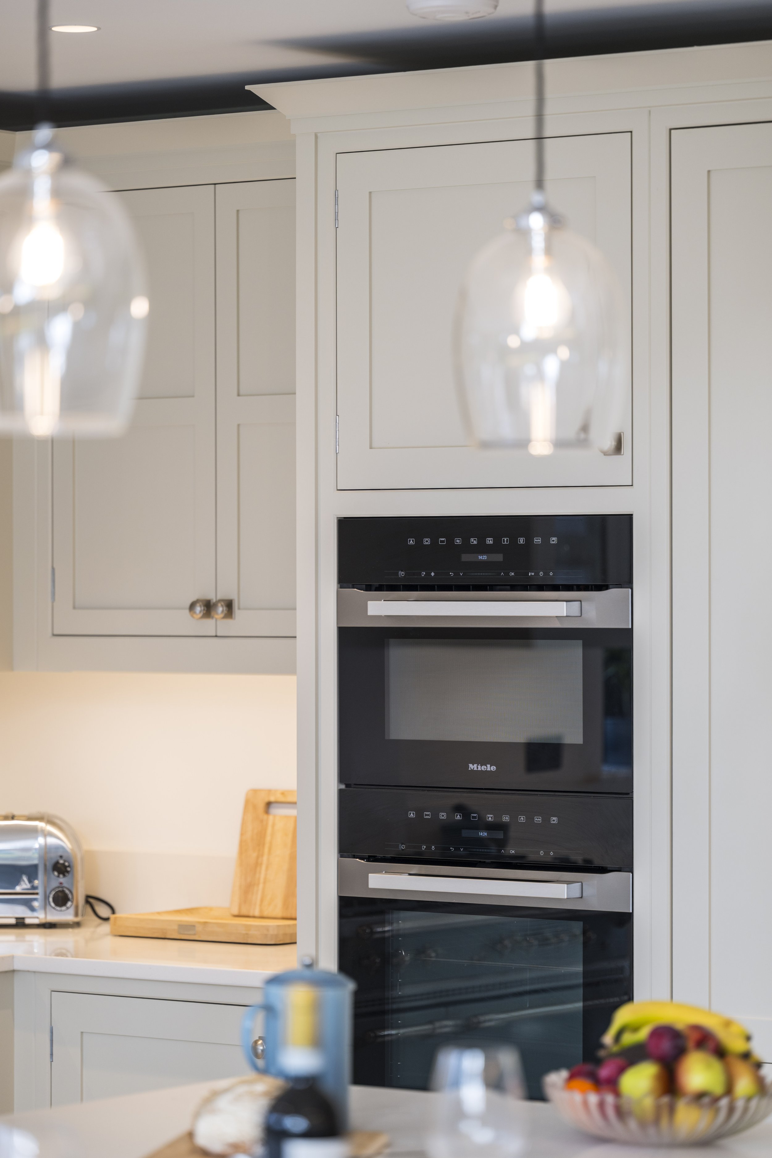 A kitchen with built-in black and stainless steel Miele oven units, white cabinetry, a toaster on the counter, and a bowl of assorted fruit in the foreground.