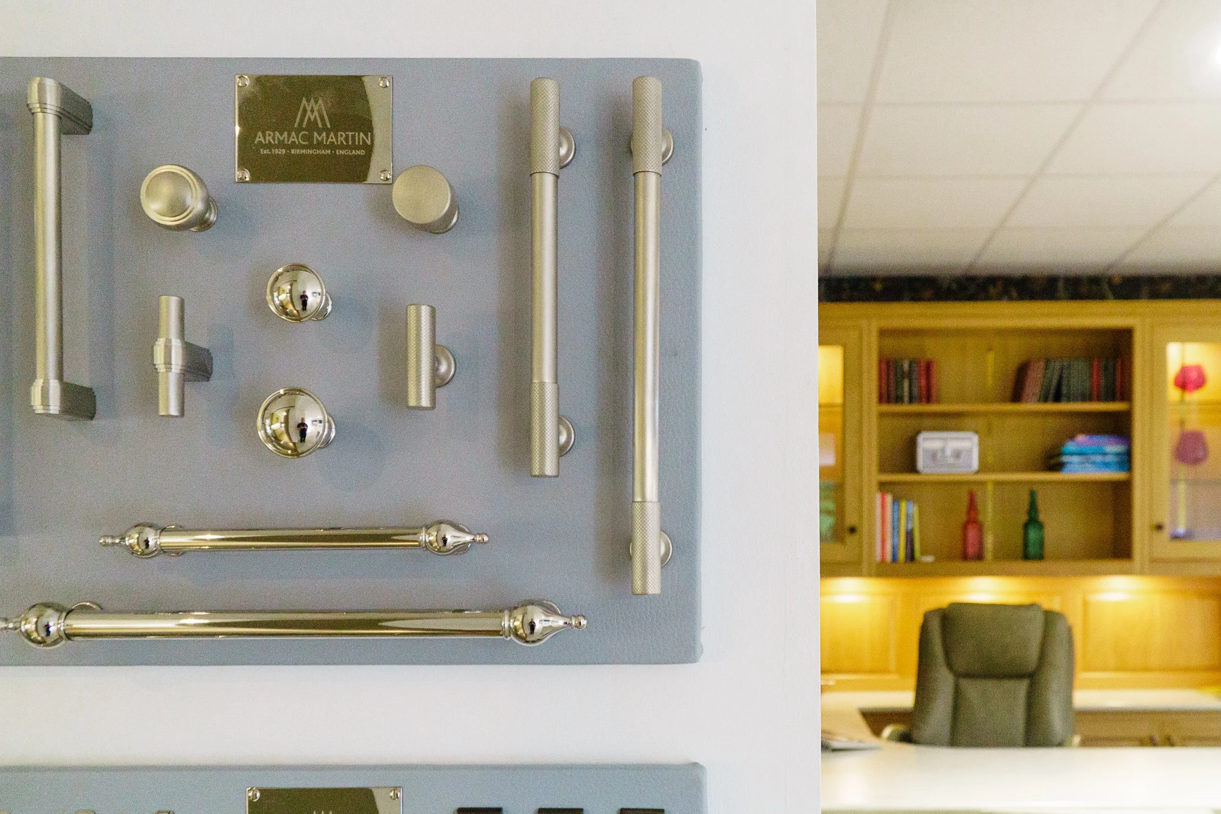 Metal door handles and hardware displayed on a blue board at a showroom, with a wooden office cabinet and bookshelf in the background.