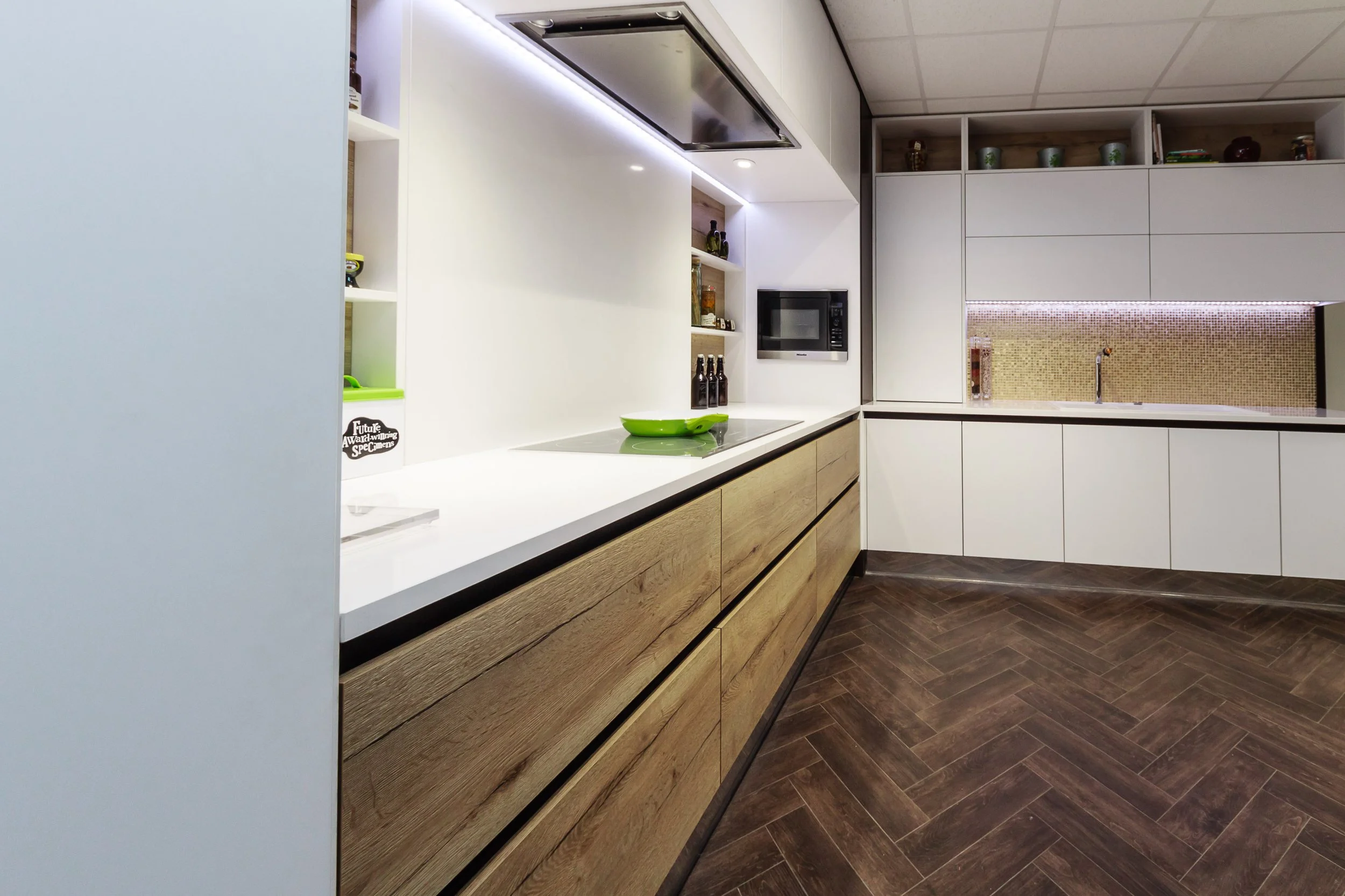 Modern kitchen with white and wood cabinetry, dark wood herringbone floor, and stainless steel appliances. Include visible countertops, a microwave, and decorative items on open shelves.