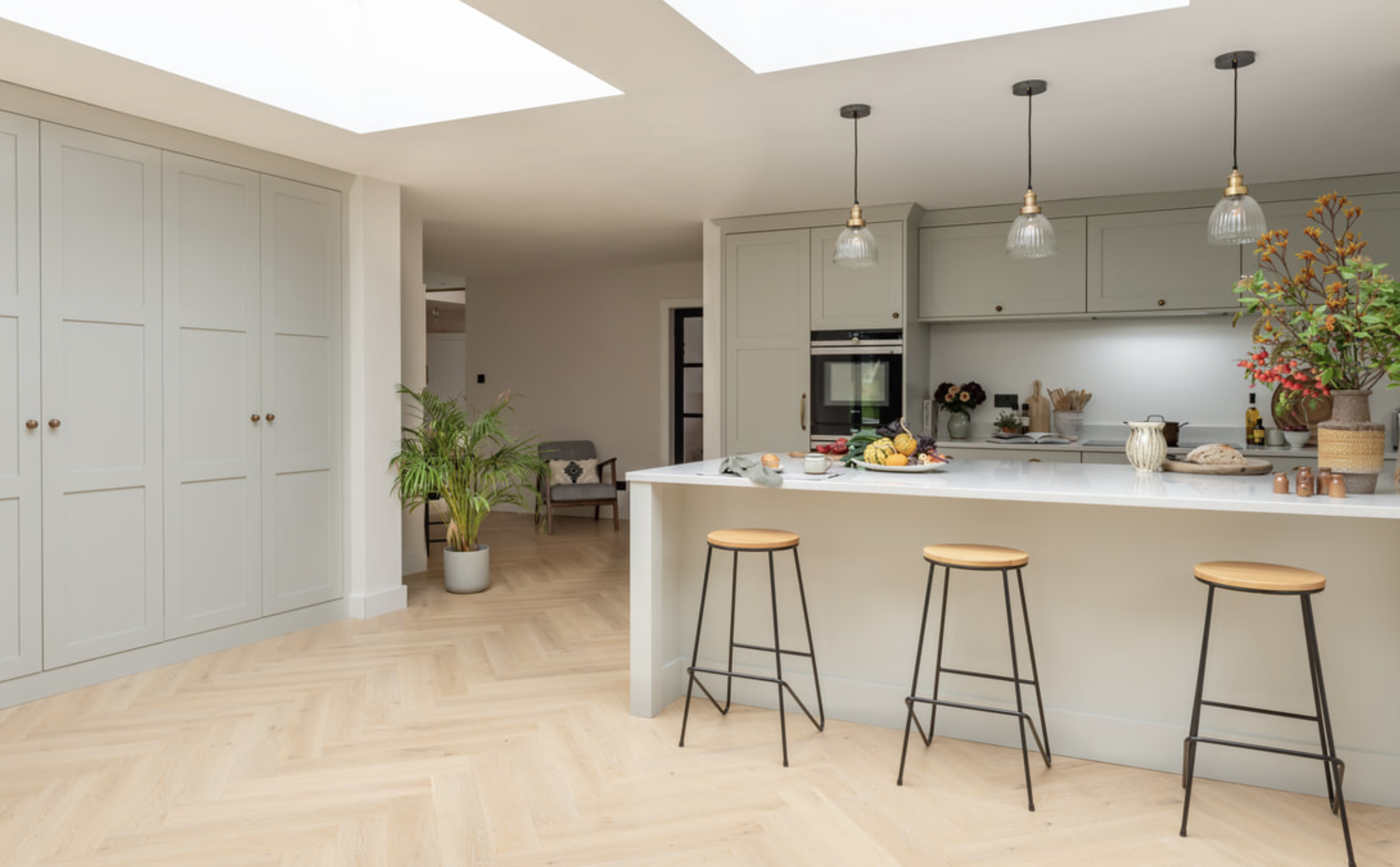 Modern kitchen with white cabinetry, a large island with three wooden stools, pendant lights, and decorative plants and vases.