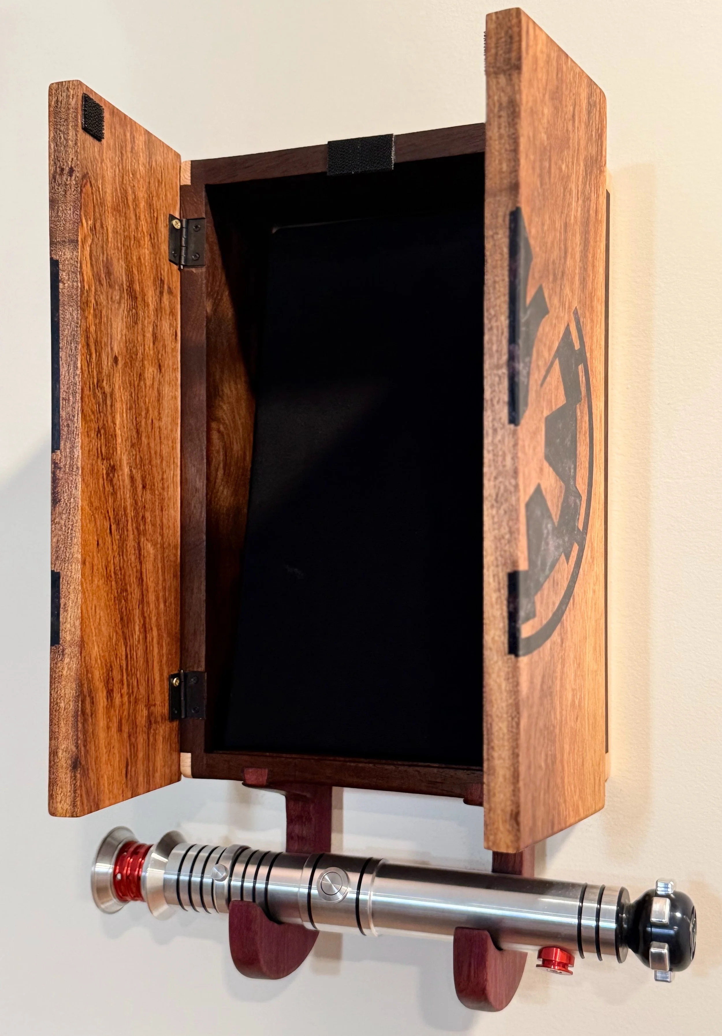 A wooden box with a black interior and a partially open lid, mounted on a wall, with a metallic lightsaber replica attached below it.