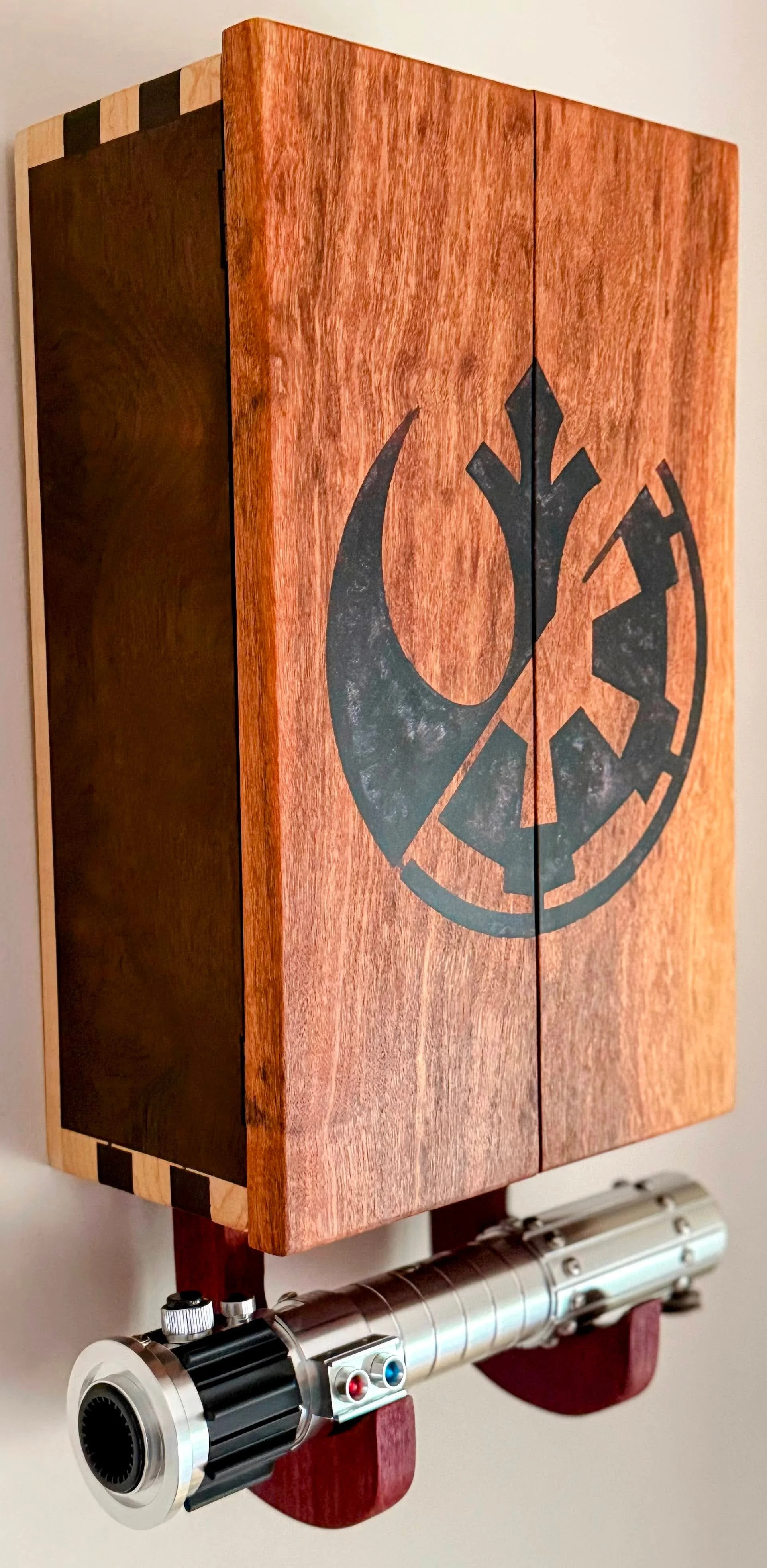A wooden wall-mounted display cabinet with a black emblem of the Galactic Empire from Star Wars on its front. Below the cabinet, there is a metallic prop lightsaber with a black grip and control buttons, mounted on the wall.