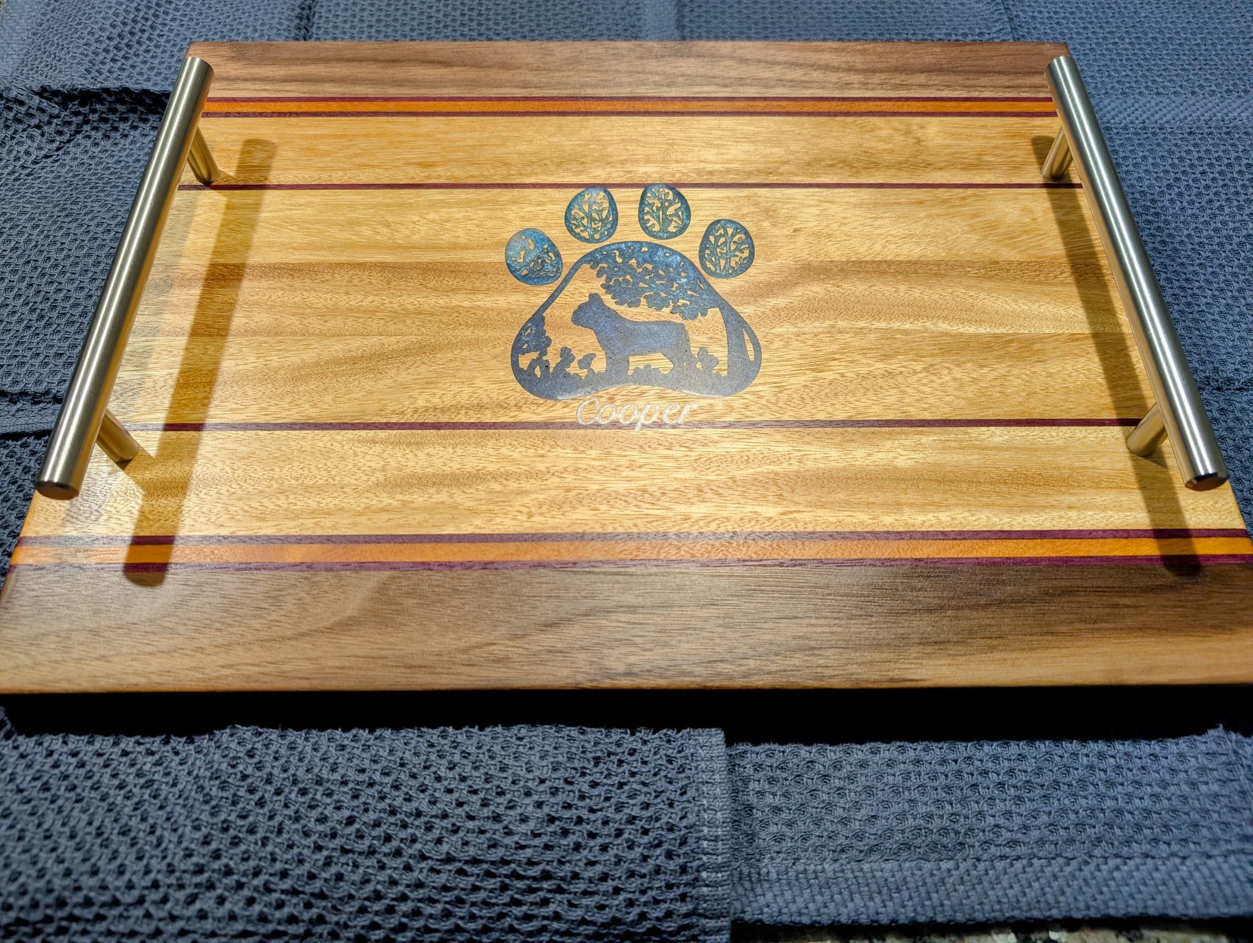 A wooden tray with metal handles, featuring a blue patch of woodland animals and trees with the word 'Cooper' underneath, placed on a dark textured fabric surface.