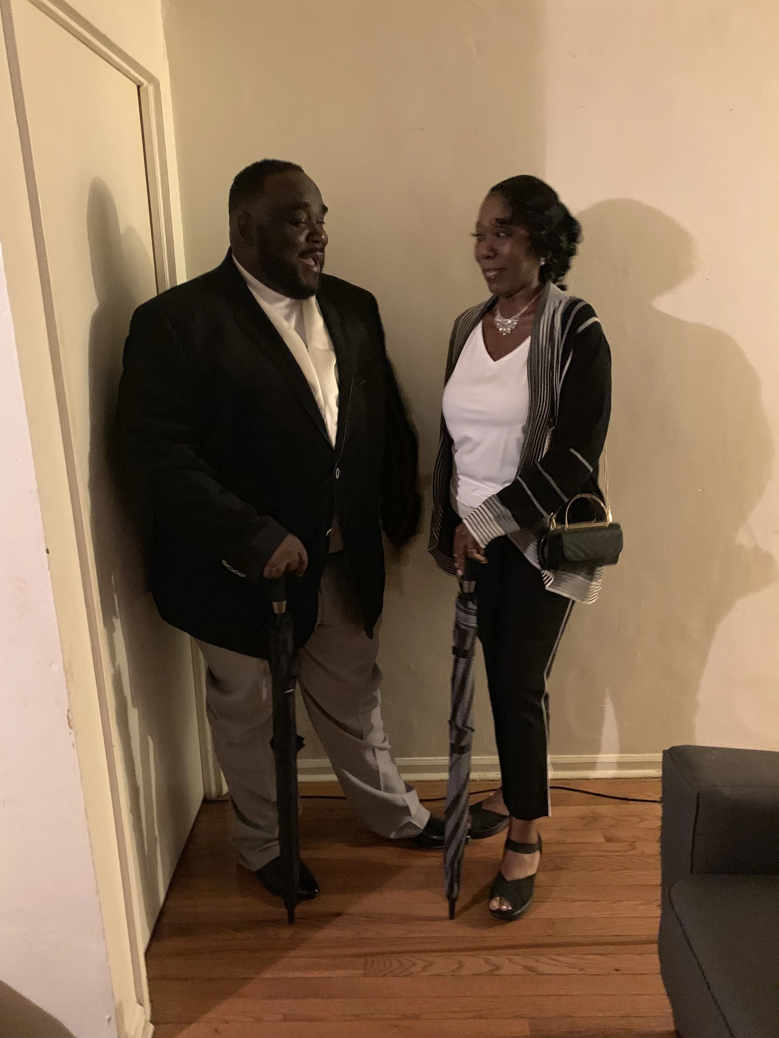 A man and woman standing together indoors, both holding umbrellas, and smiling at each other.