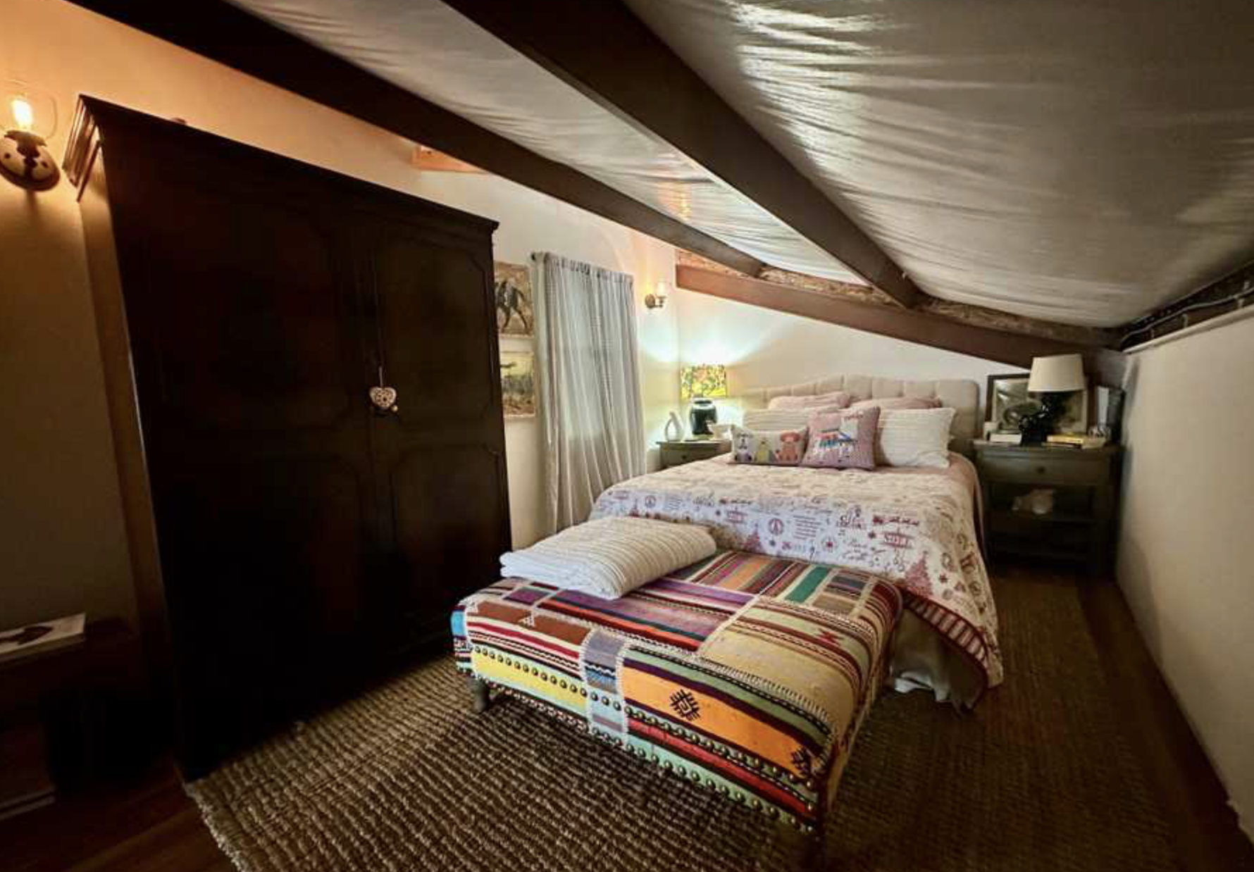 Cozy bedroom with sloped ceiling, dark wooden wardrobe, and a bed with pink and white bedding, colorful pillows, and a matching bench at the foot of the bed. There's a nightstand with a lamp and decorative items, and a window with white curtains.