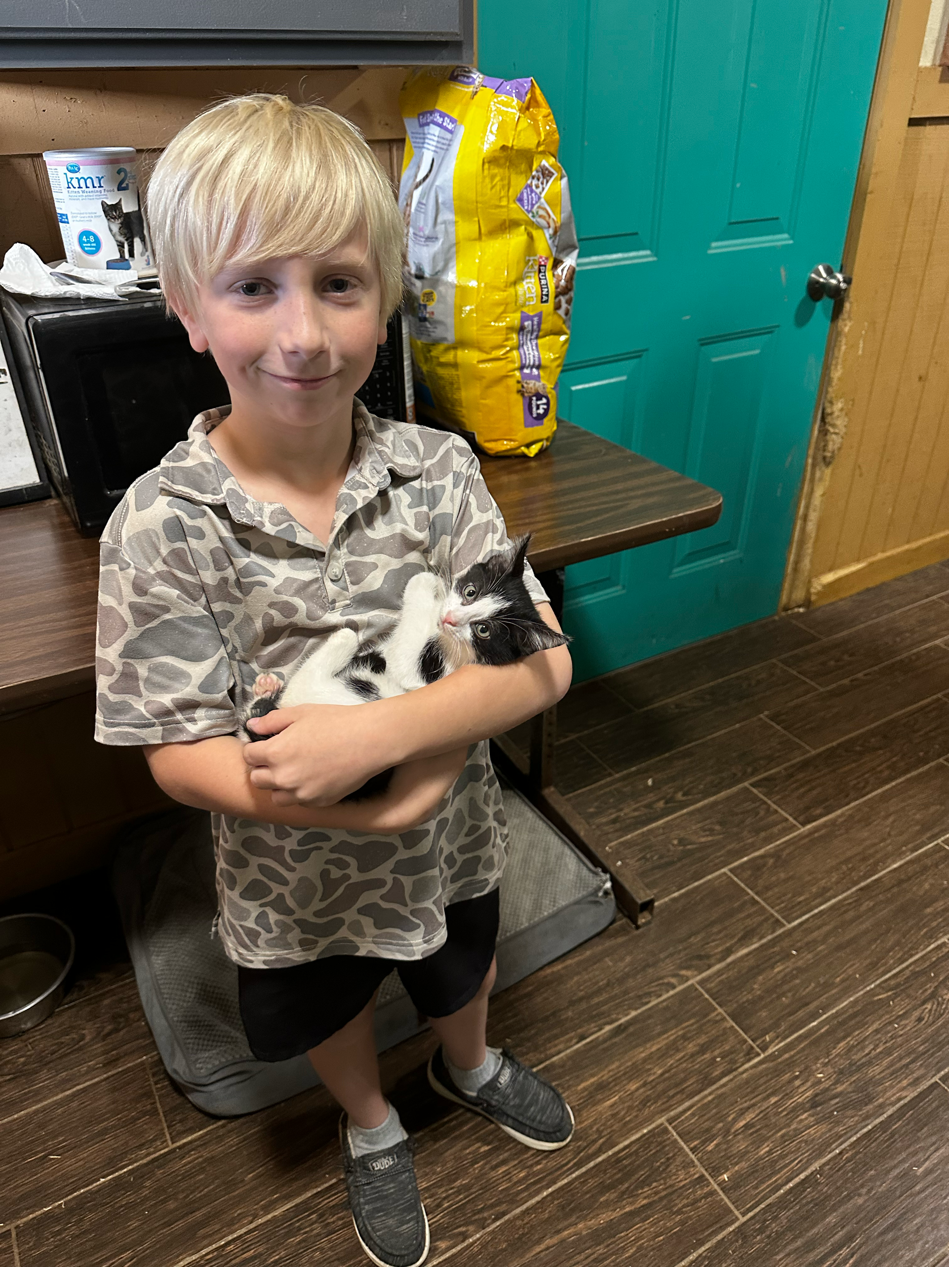 A young boy with blond hair holding a black and white kitten in a kitchen. The boy is wearing a leopard print shirt and black shorts, standing on a wooden floor. In the background, there is a teal door, a bag of cat food, a microwave, and some paper 