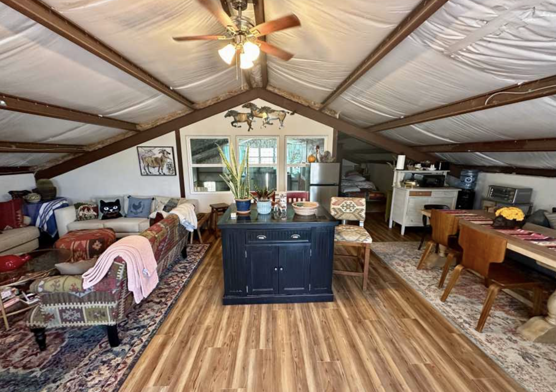 Interior view of a living room with sloped ceiling, ceiling fan, couch with pillows, and a dining table with chairs, featuring wooden floors and various decorative items.