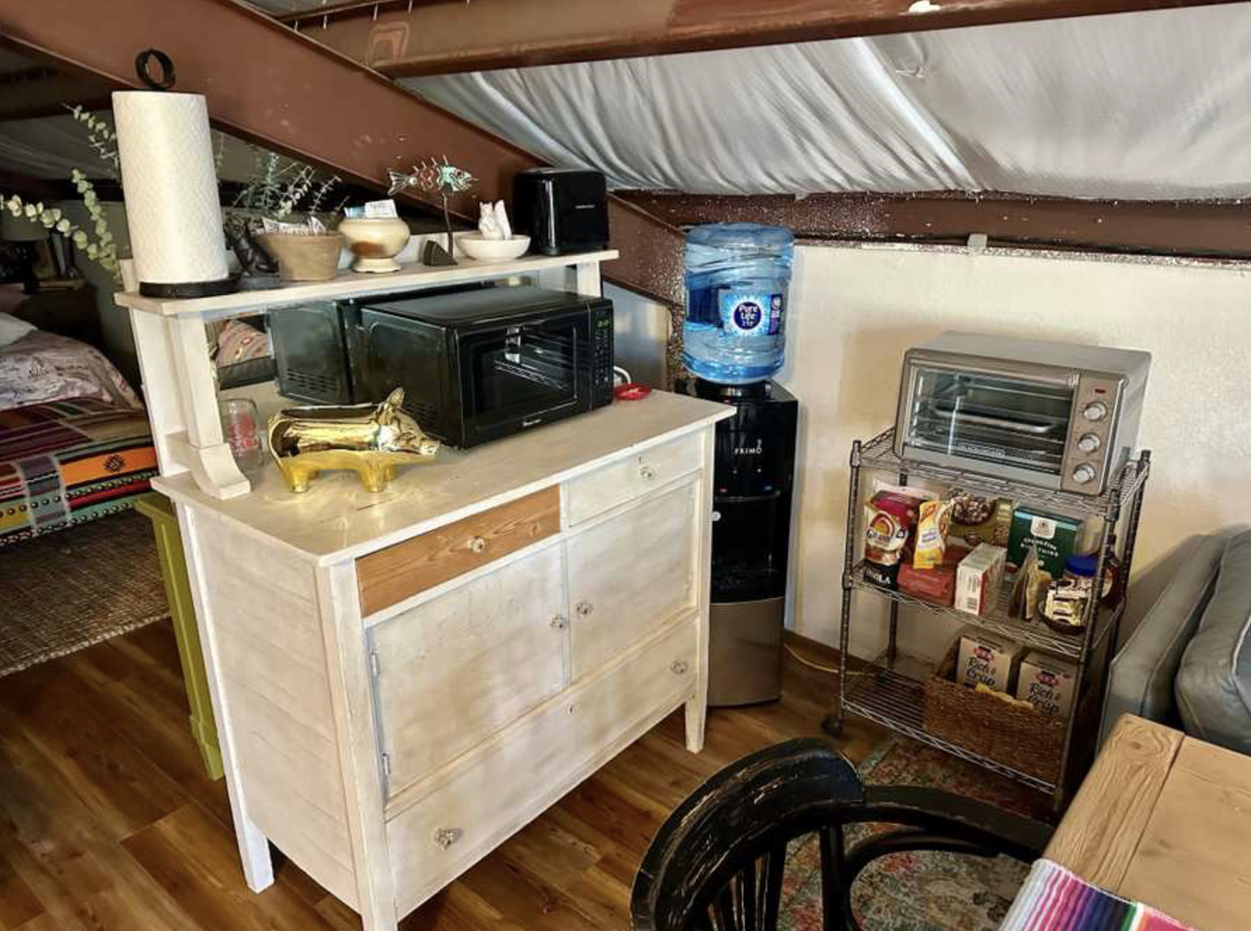 Small kitchenette with microwave and water cooler, surrounded by various kitchen items and a shelf with snacks, in an attic space with sloped ceiling.