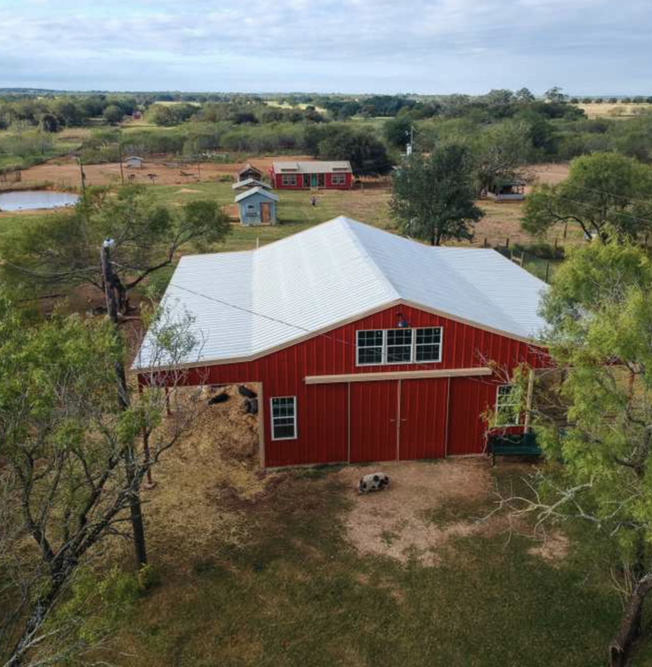 A red barn with a white metal roof in a rural setting, surrounded by trees and open land, with small additional buildings in the background.