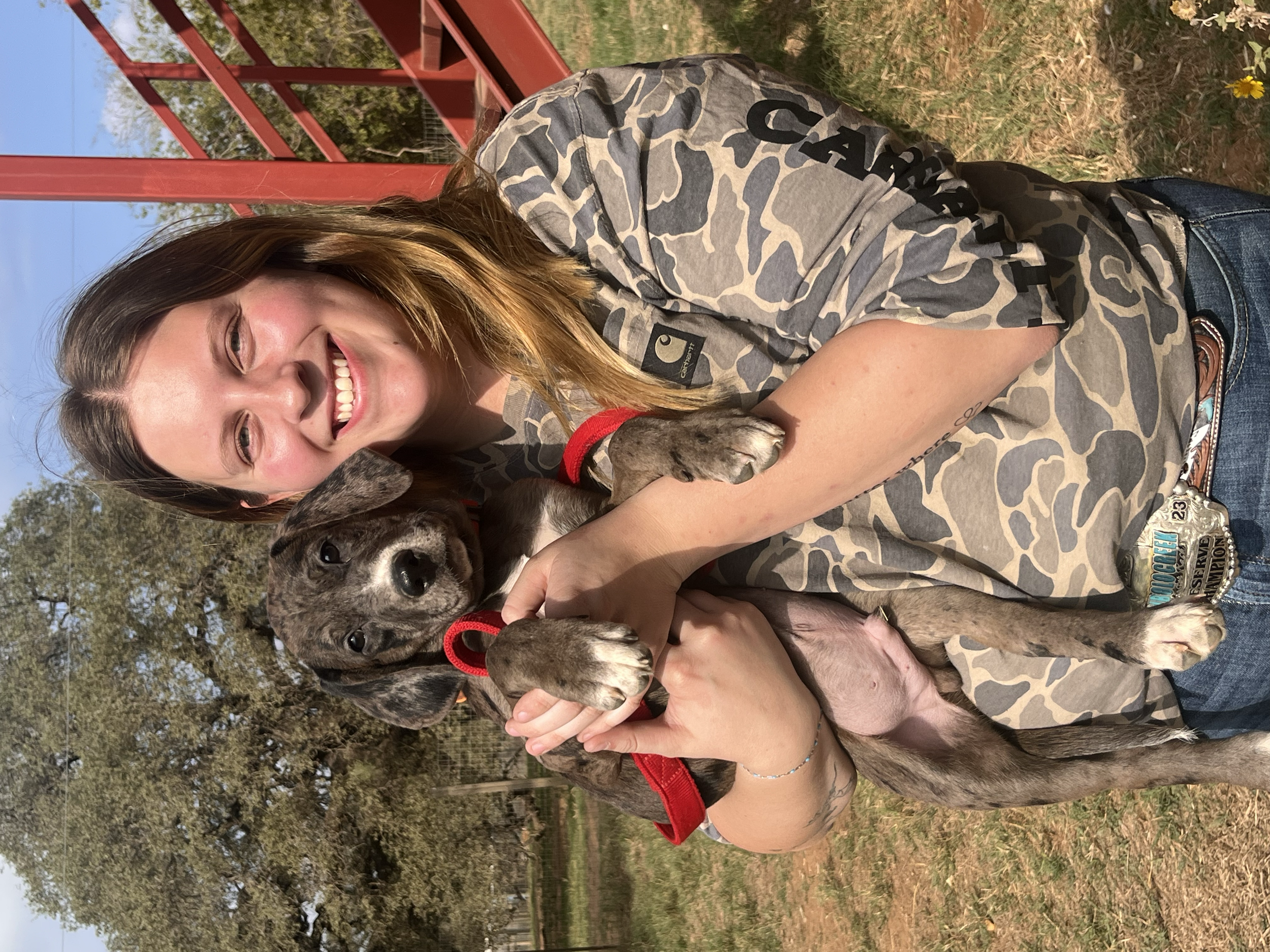 A woman with long brown hair smiling while holding a puppy with brindle coat and long ears, outdoors on grass under a blue sky.