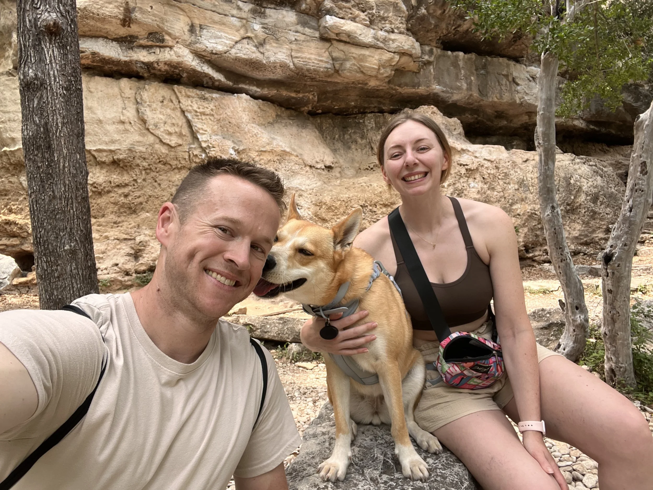 A smiling man and woman taking a selfie outdoors with their dog, sitting on a rock near trees and a rocky background.