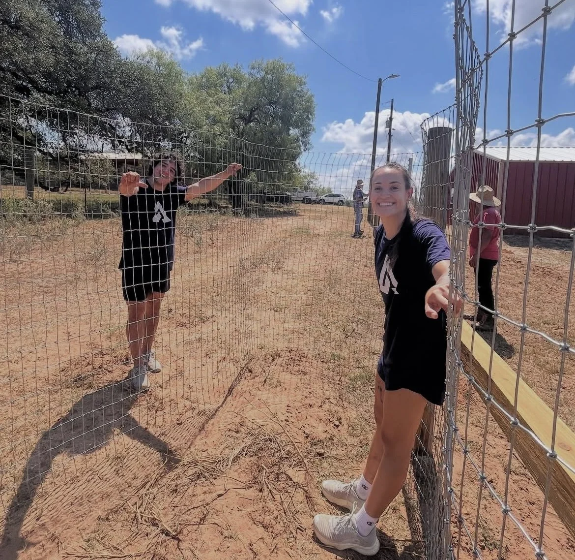 Two young women smiling and leaning on a metal fence in a rural outdoor setting on a sunny day, with a few other people in the background and trees in the distance.