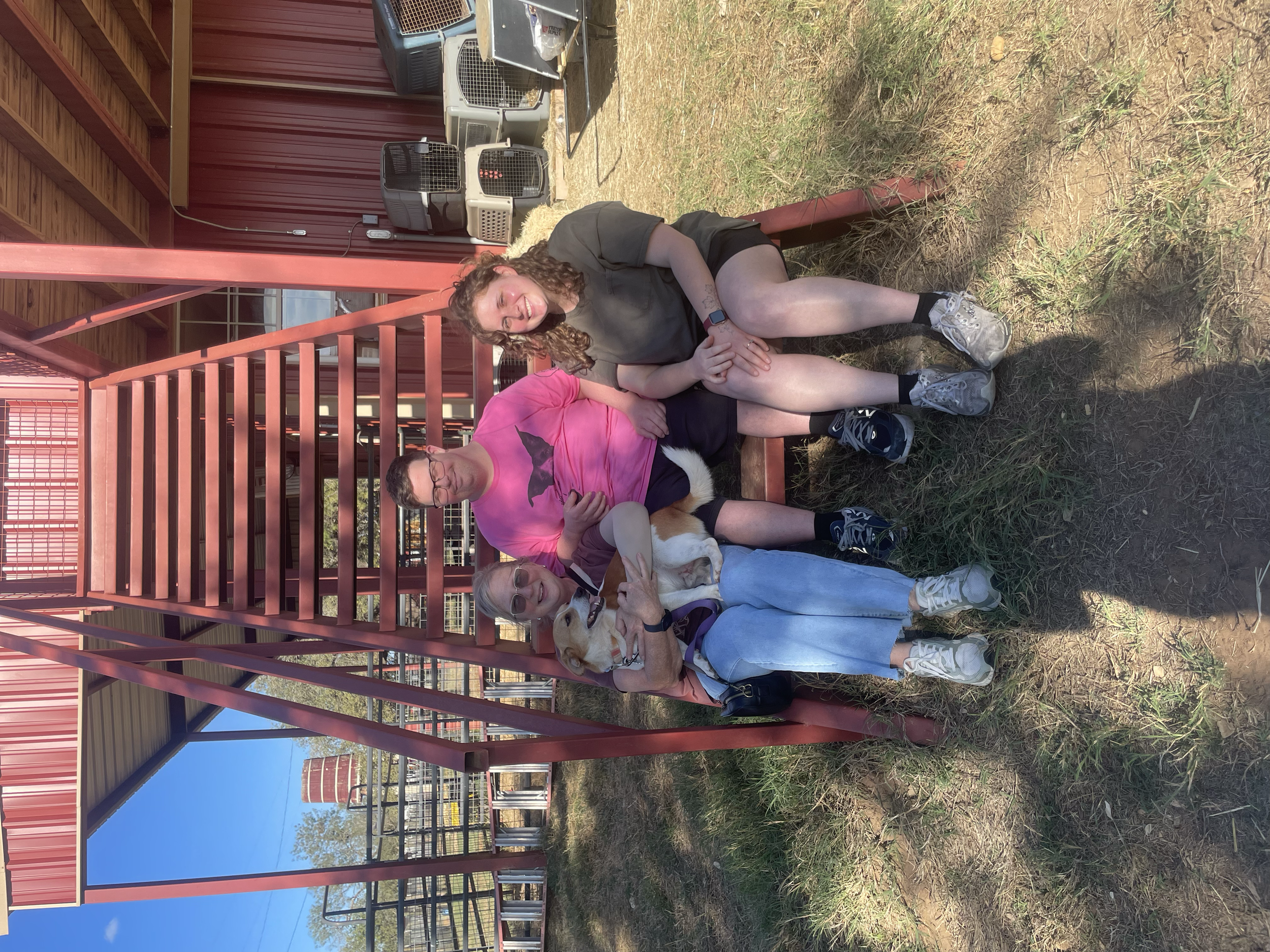 Three women and a dog sitting on a red metal bench under a red metal staircase at a pet adoption event or animal shelter. The women are smiling, and the dog is a white and tan mixed breed with a purple harness.