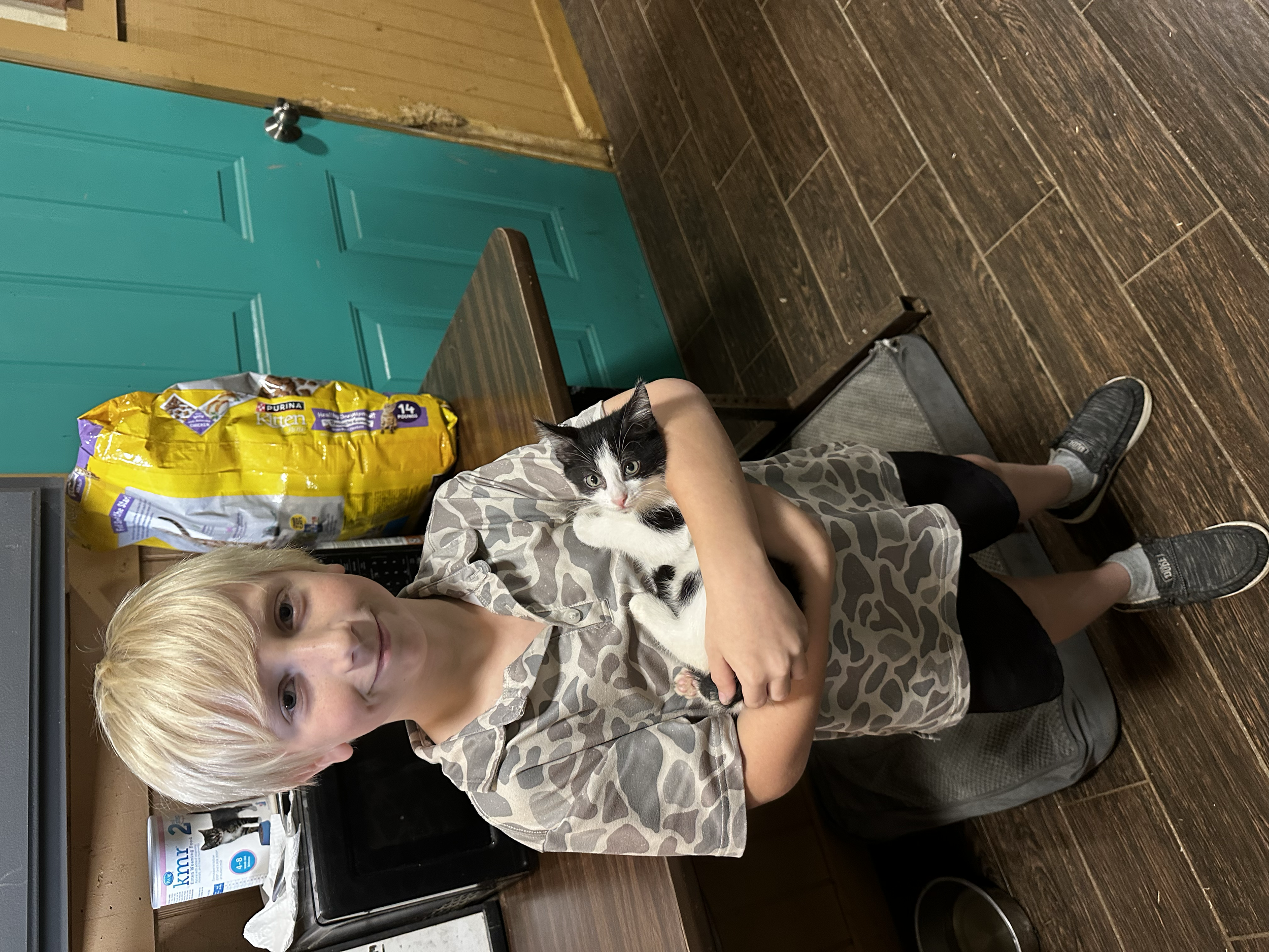 A young boy with blond hair holding a black and white kitten in a kitchen. The boy is wearing a leopard print shirt and black shorts, standing on a wooden floor. In the background, there is a teal door, a bag of cat food, a microwave, and some paper 