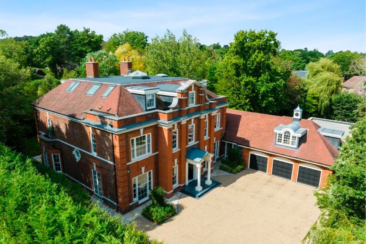 A large red brick mansion with a tiled roof, multiple chimneys, and a three-car garage, surrounded by green trees on a sunny day.