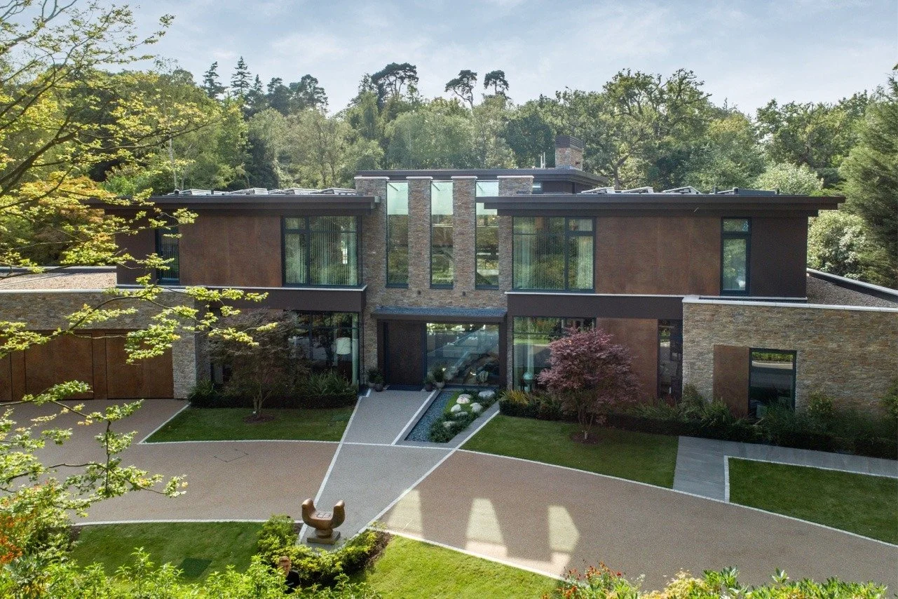 A modern two-story house with large windows, brick and wood siding, surrounded by green trees and landscaped yard with walking paths and plants.