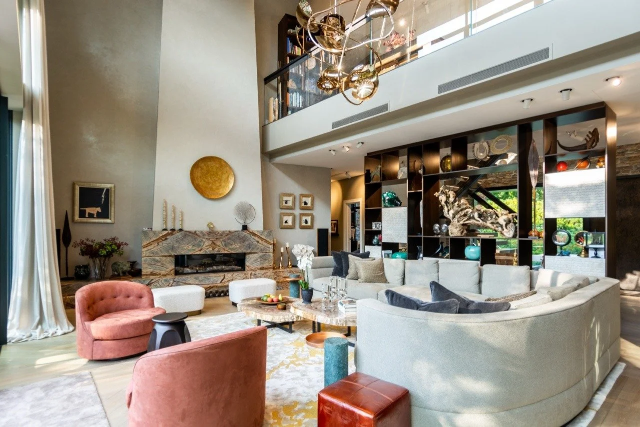 Spacious modern living room with a curved cream sofa, pink chairs, a marble coffee table, a fireplace, and a built-in bookshelf with decorative items, artwork, and large windows providing natural light.