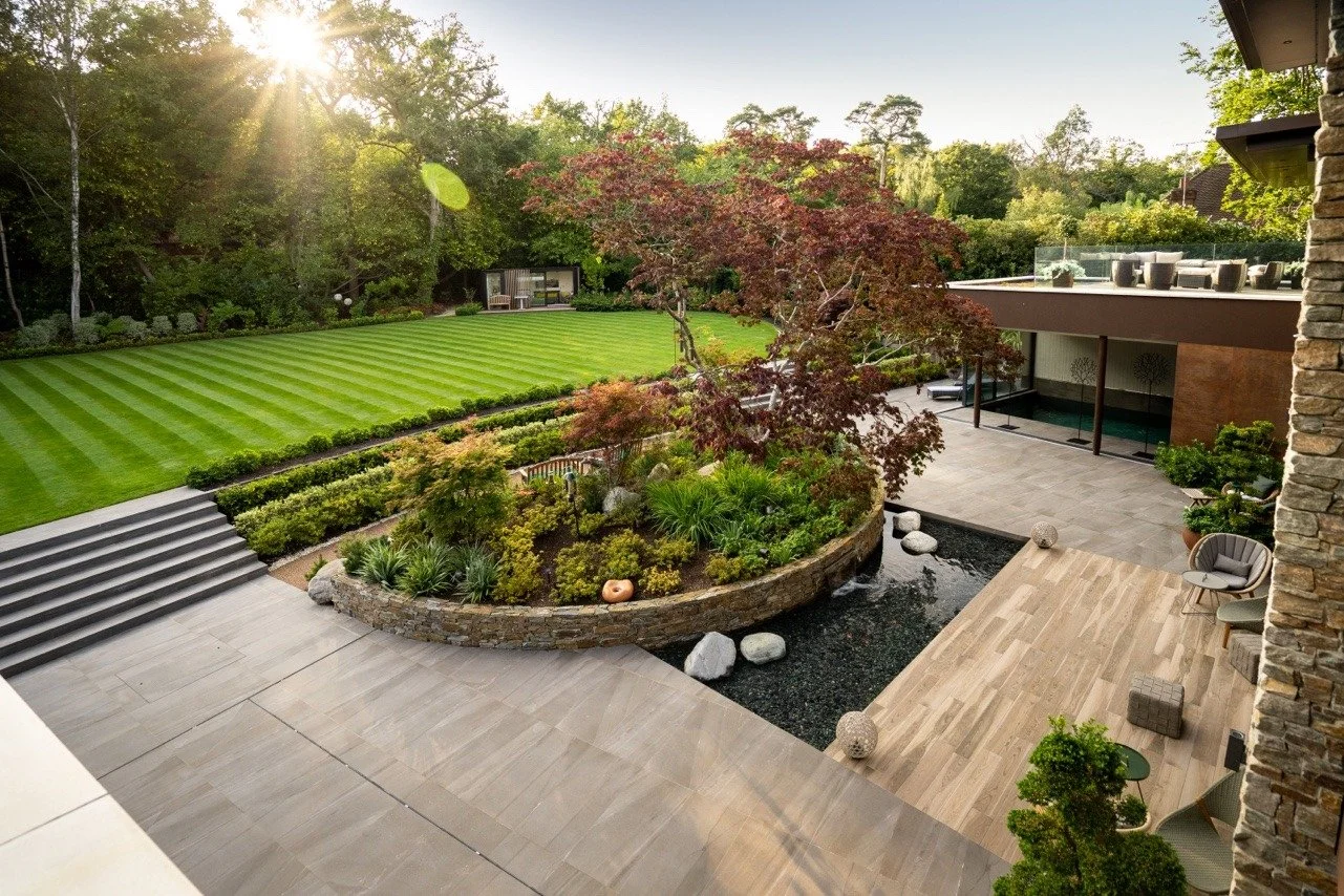 Luxury backyard with a well-maintained lawn, landscaped garden, a small pond with rocks, and shaded seating area on a patio, surrounded by trees and modern house architecture.