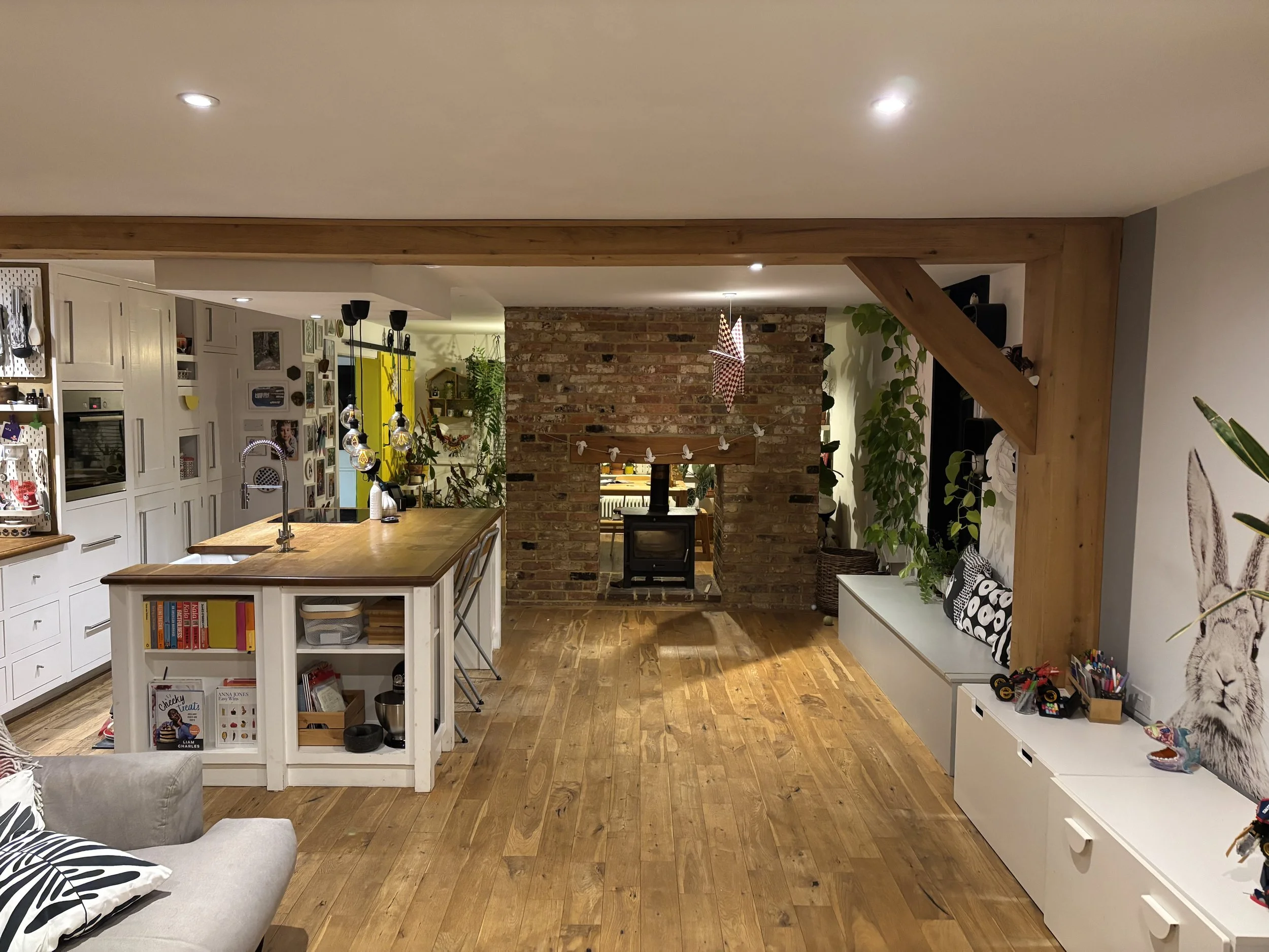 A cozy open-plan living space with a brick fireplace, wooden flooring, and a kitchen island with pendant lighting. There is a gray sofa with black and white pillows, a built-in bench with cushions, and decorative plants.