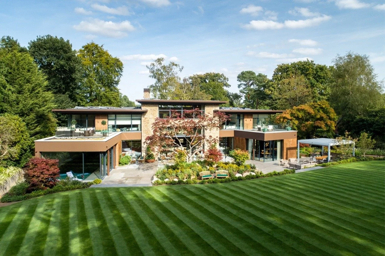 Modern house with large glass windows and a flat roof, surrounded by a well-maintained lawn and lush trees, under a partly cloudy sky.