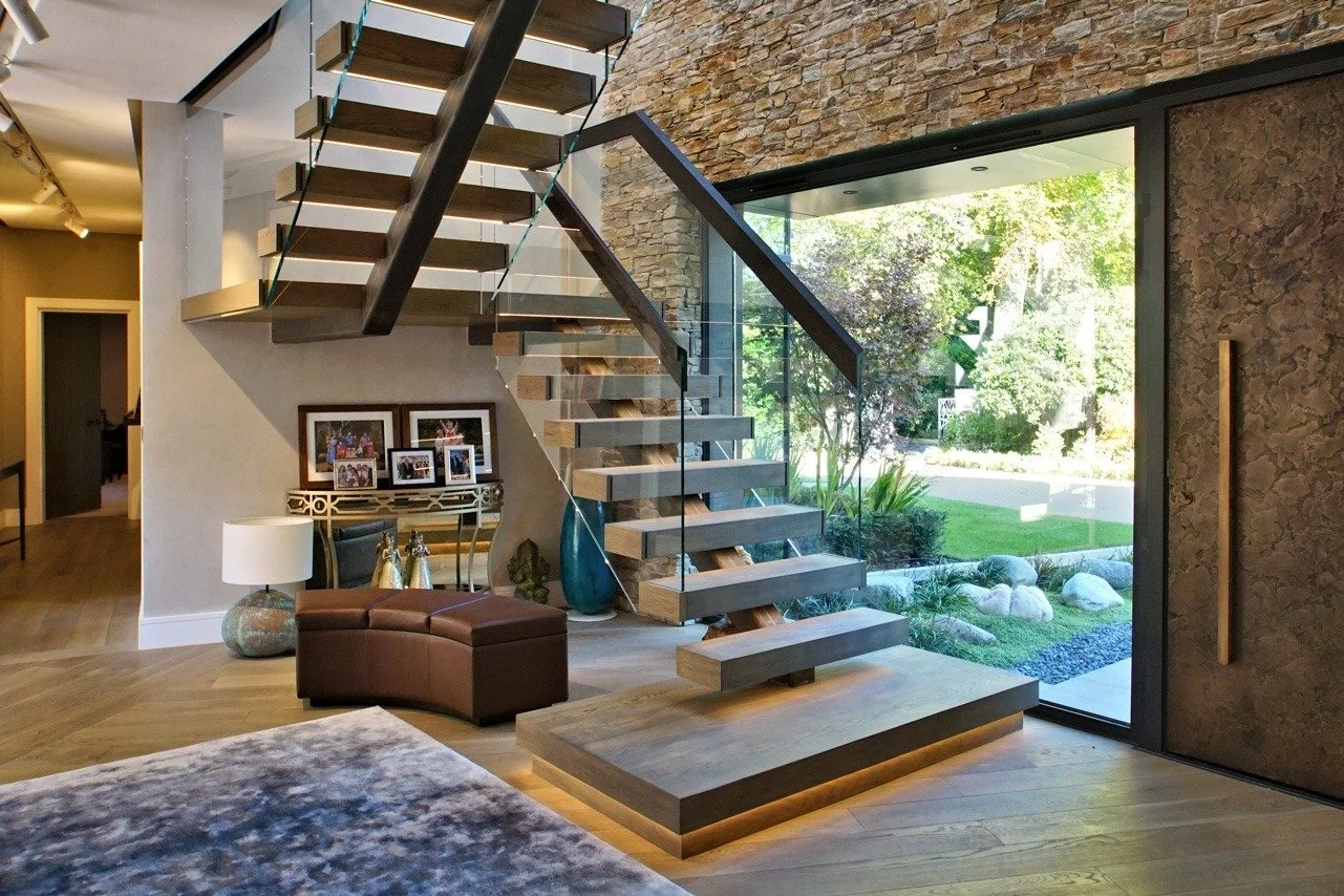 Modern interior featuring a floating staircase with open wooden steps, glass railings, a stone accent wall, large glass window showing greenery outside, a leather ottoman, a table with family photos, a table lamp, and decorative pottery.