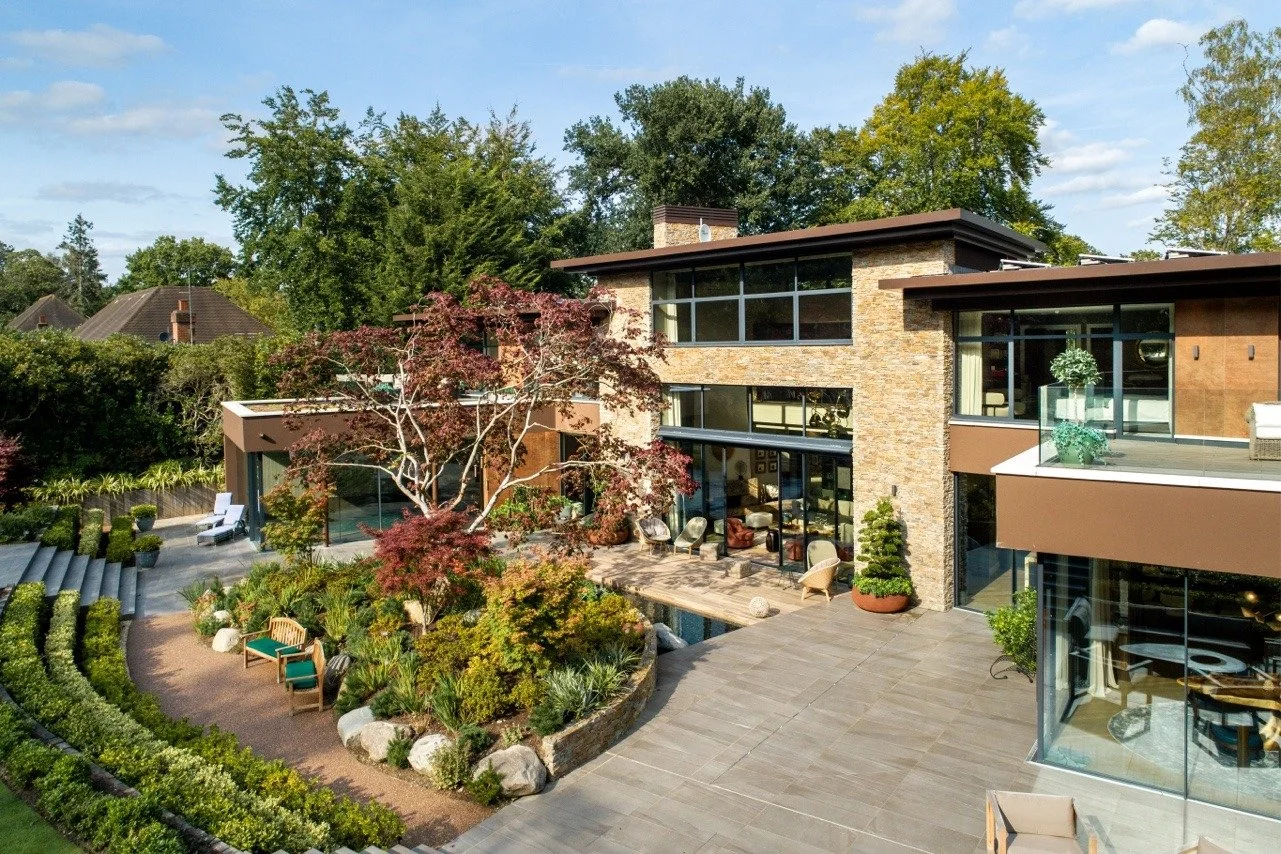 Modern multi-story house with large glass windows, surrounded by well-maintained gardens and outdoor seating areas, under a partly cloudy sky.