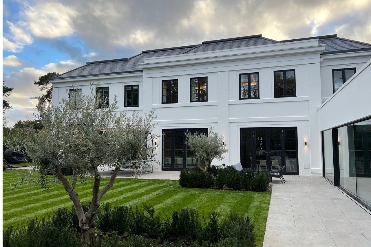 Modern white two-story house with large black window frames, a lush green lawn, a paved patio, and a cloudy sky above.