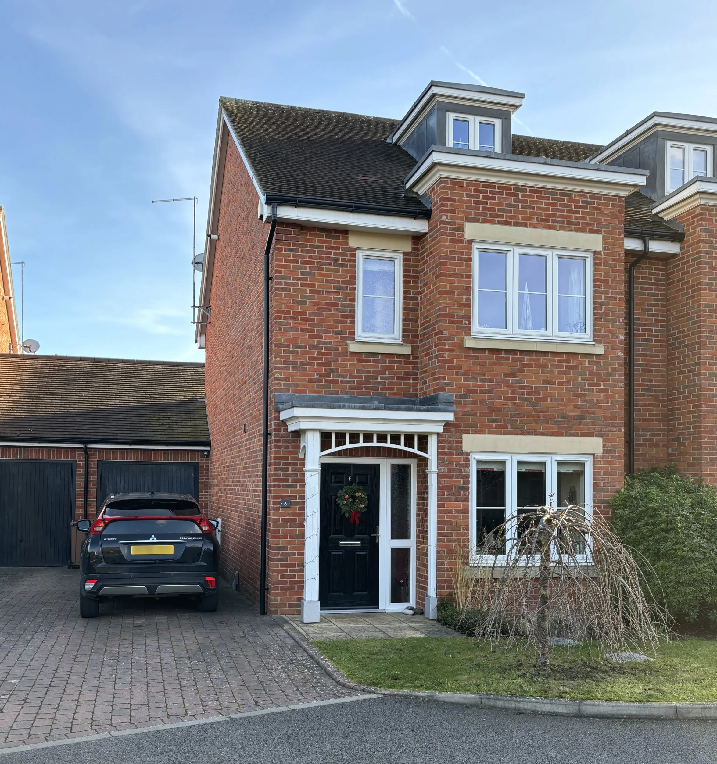 A modern brick townhouse with a black front door decorated with a festive wreath, a small lawn, a leafless tree, and a parked black SUV on the driveway, under a clear blue sky.
