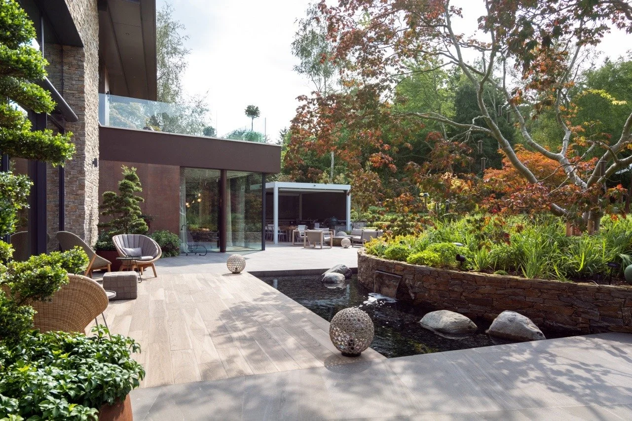 Modern outdoor patio with seating, decorative balls, and a water feature, surrounded by lush trees and greenery.