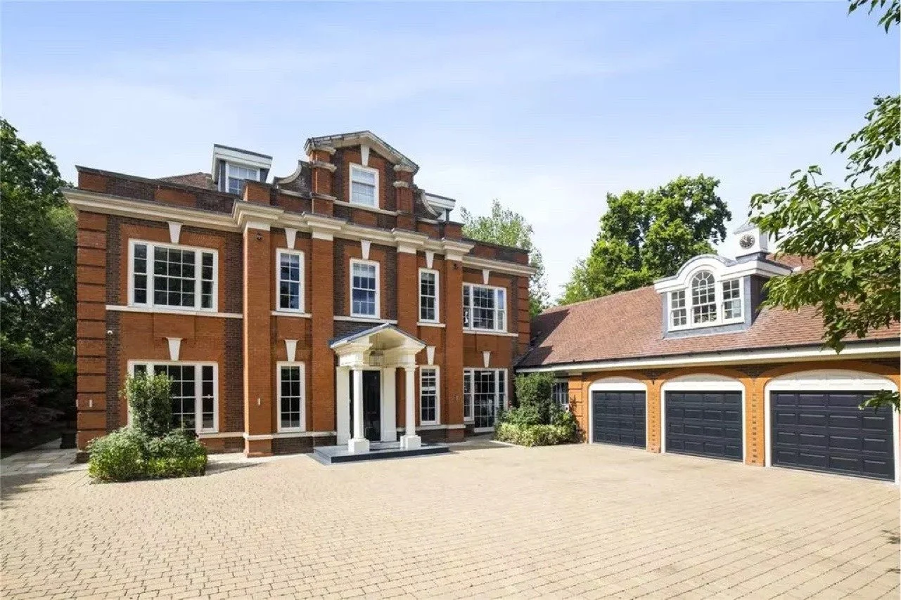 A large brick house with white accents and multiple windows, attached to a garage with three black doors and a red-tiled roof, surrounded by trees.
