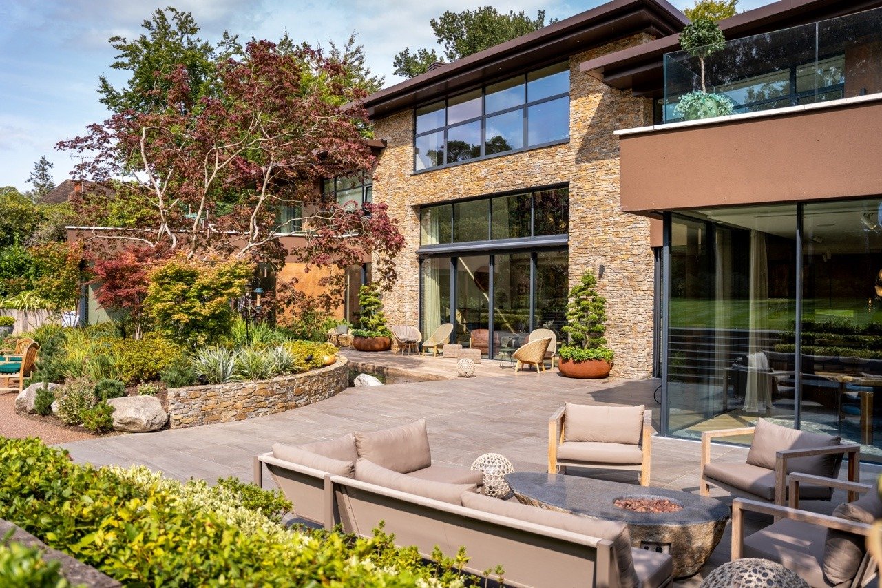 Modern house with large glass windows, stone facade, and outdoor patio area with chairs, sofas, and a fire pit, surrounded by lush landscaping and trees.