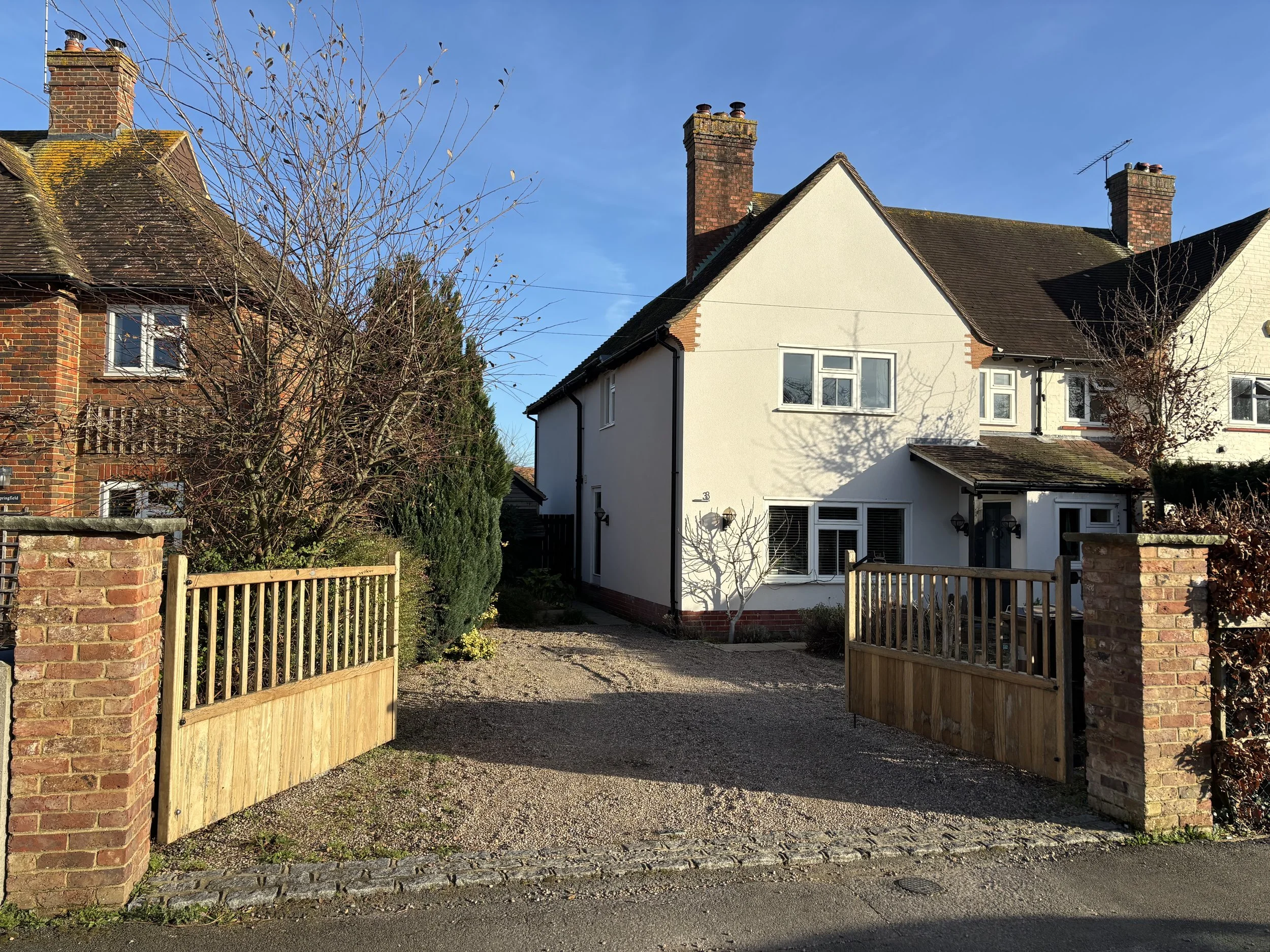 Two houses with brick and white exteriors are separated by a driveway with a wooden gate. Several leafless trees and bushes are in the front yard and the sky is clear and blue.