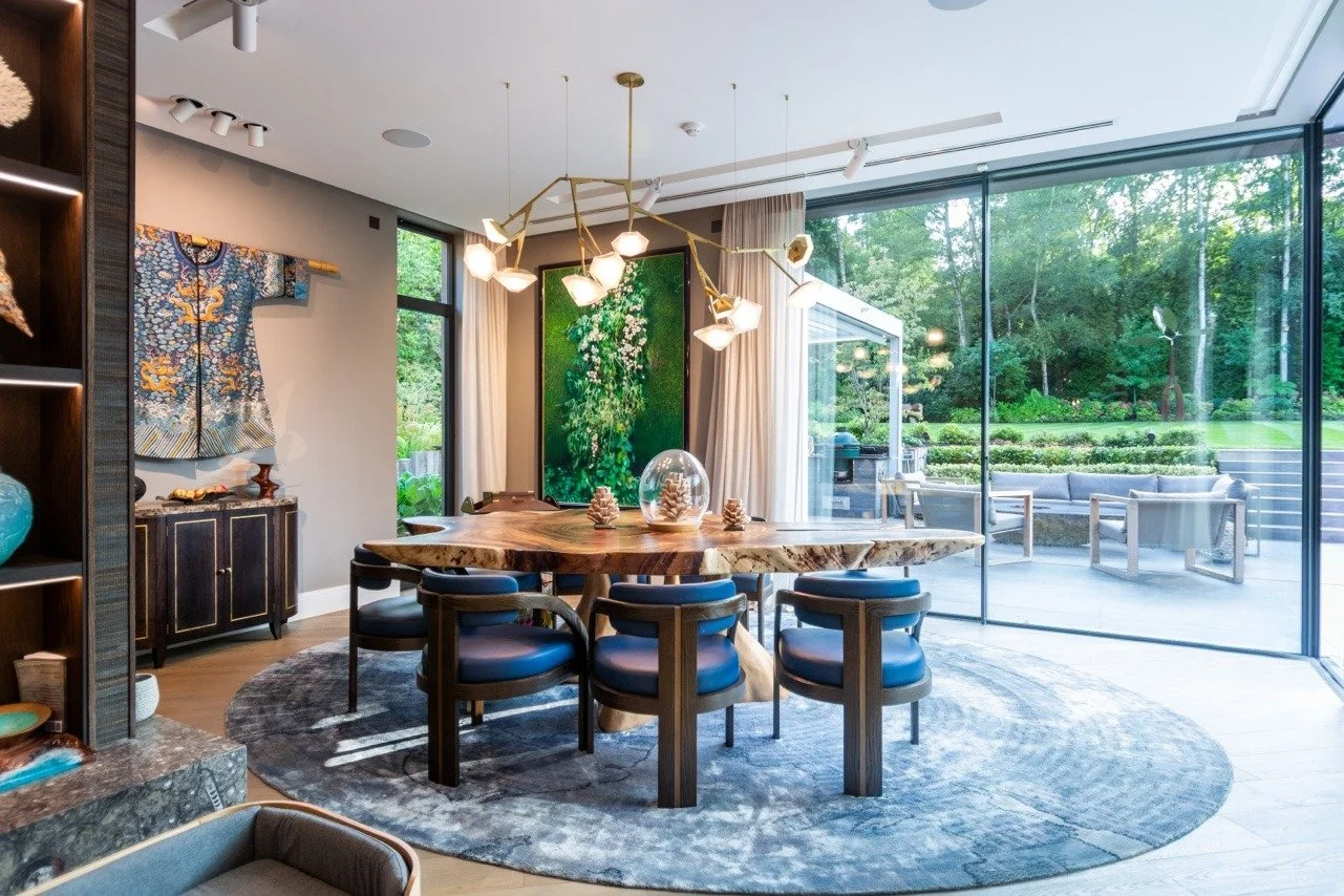Modern dining room with large wooden table, six chairs with dark frames and blue cushions, a circular rug, and a chandelier with multiple geometric light fixtures. Large glass sliding doors provide a view of a patio with outdoor seating and a green l