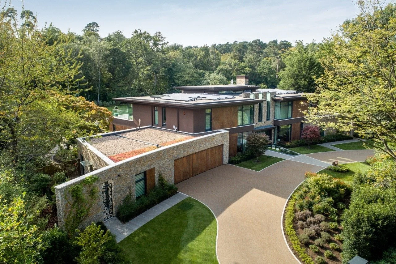 A modern house surrounded by trees with a curved driveway leading to a garage and landscaped yard.