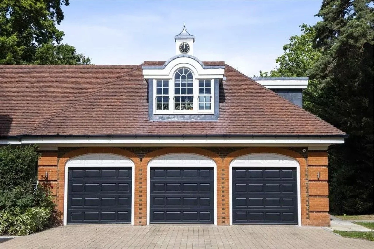 A brick building with a three-car garage with black doors, a large arched window with white framing above the garage, and a small clock tower on the roof with a clock showing 4:05. There are trees on either side of the building and a paved driveway i