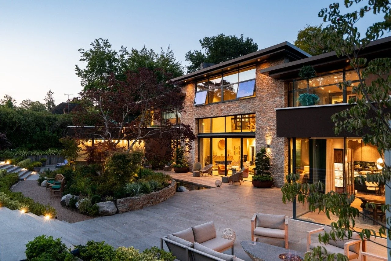 Modern house exterior at dusk with well-lit interior, large glass windows, stone accents, and outdoor seating area surrounded by greenery.