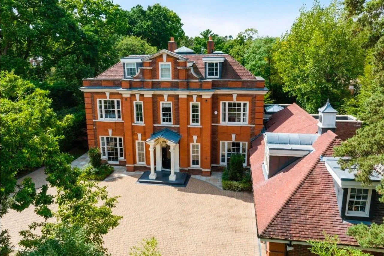 A large, ornate, three-story brick house with white accents, a front porch with columns, and a red tile roof, surrounded by lush green trees.