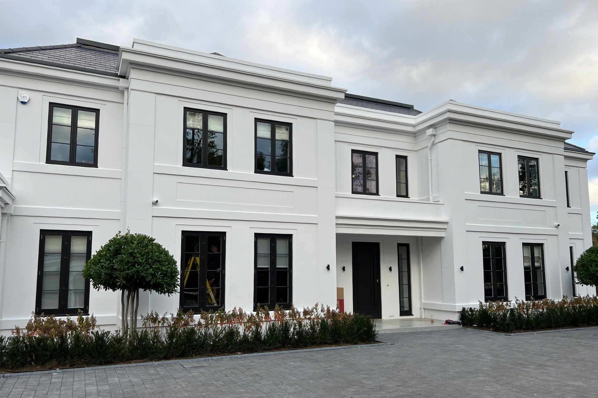 Front view of a white, multi-story house with black window frames and a black front door. The house has landscaped bushes and a paved driveway.