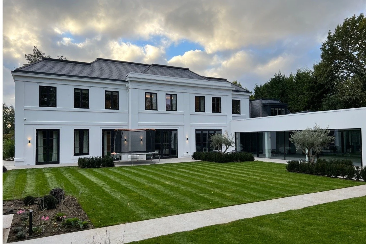 Modern white multi-story house with large windows, a well-maintained lawn with parking stripes, and a circular driveway, under a partly cloudy sky.