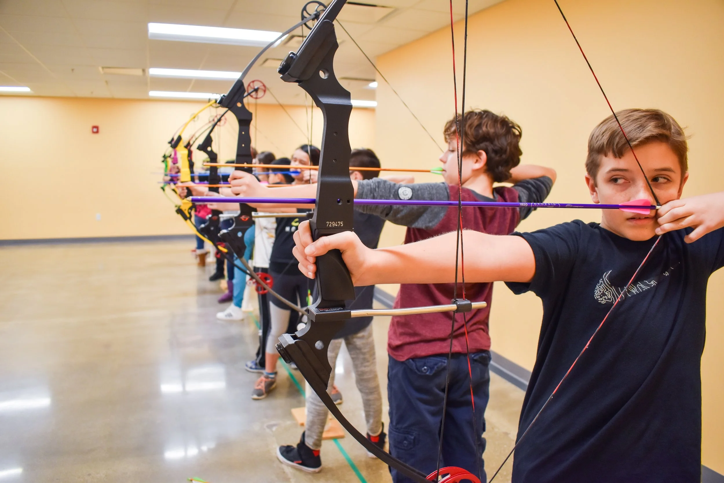 A group of children practicing archery indoors, lined up and aiming their bows with arrows ready to shoot.