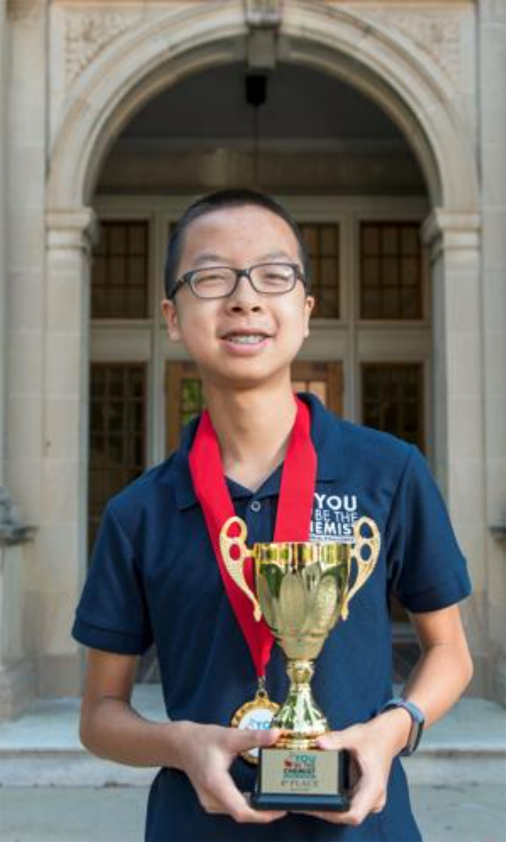 A young man with glasses smiling, wearing a navy polo shirt, holding a gold trophy with a red and gold medal hanging around his neck, standing in front of a historic building with arched entrance.