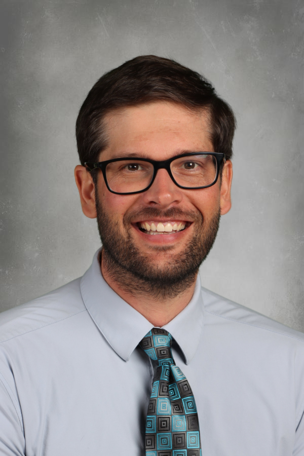Professional headshot of a man with brown hair and beard, wearing glasses, a light blue collared shirt, and a patterned tie, smiling against a gray background.