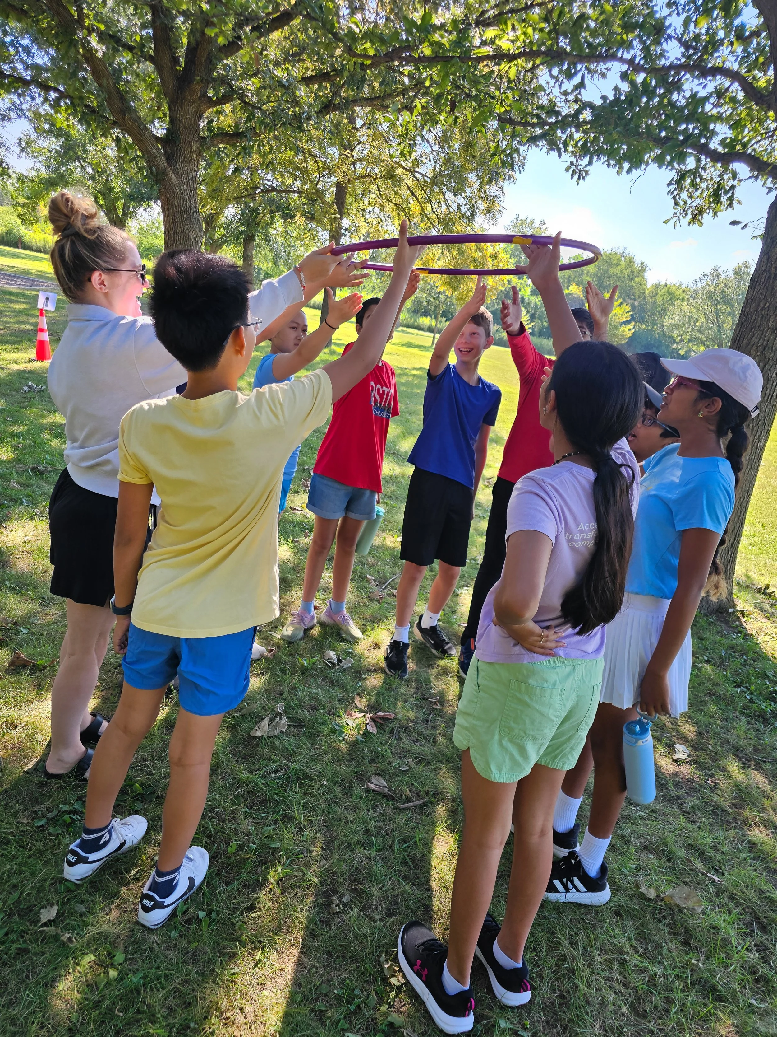 A group of children and two adults gather outdoors under a tree, playing with a purple hula hoop.