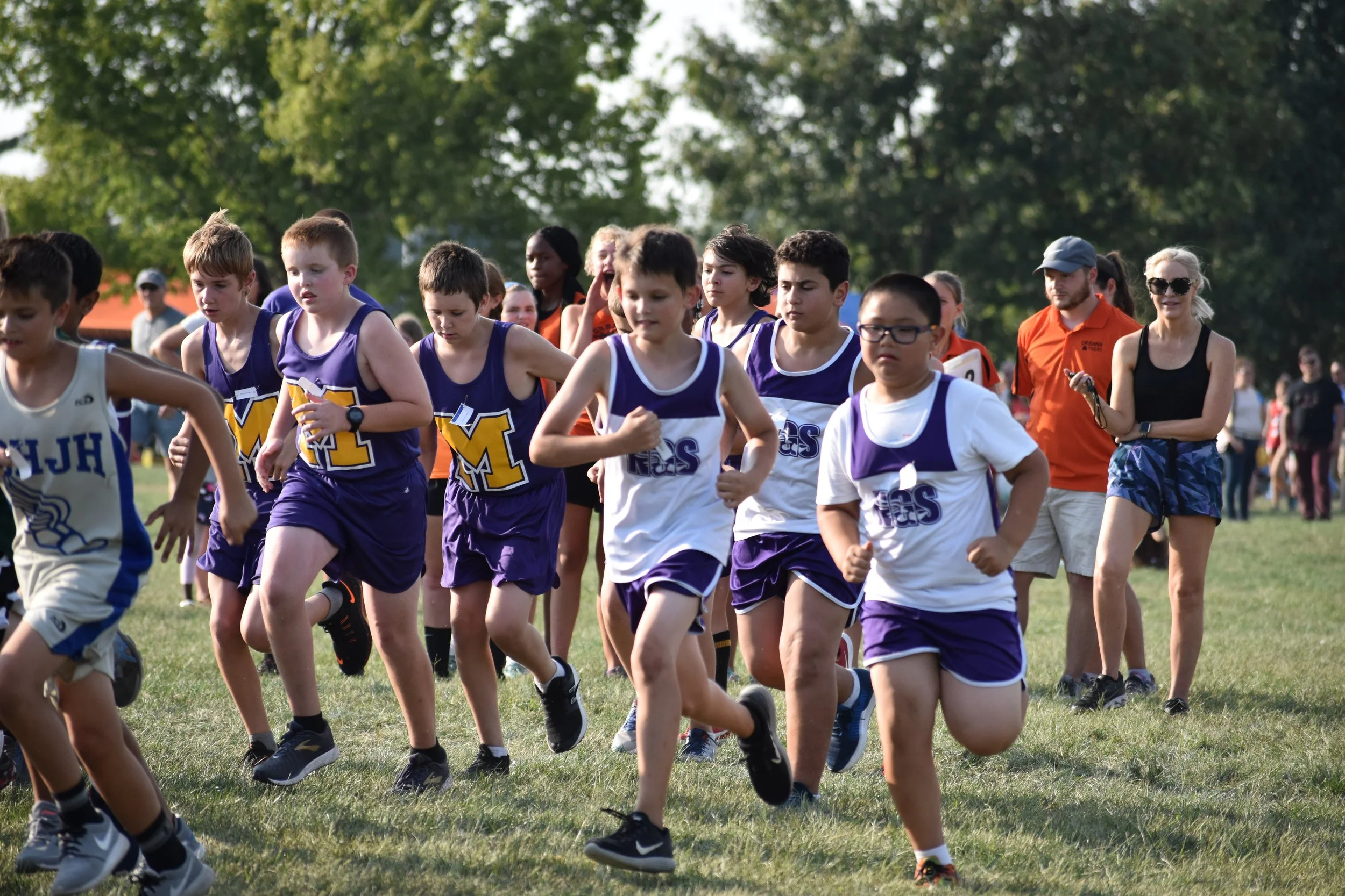Children participating in an outdoor running race on a grassy field, with spectators and coaches watching in the background.
