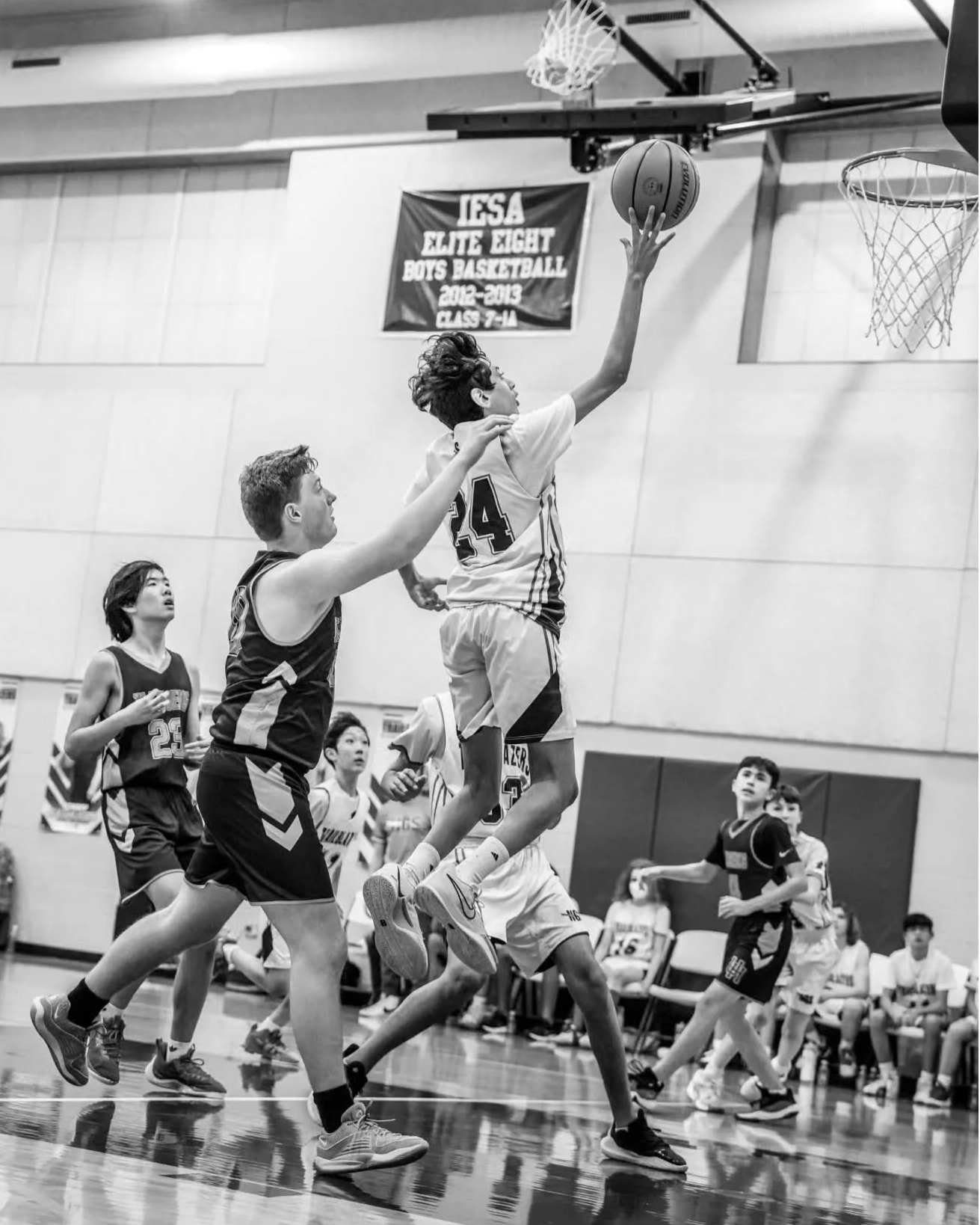 Young basketball player jumping to shoot the ball near the hoop during a game, with other players and spectators watching.