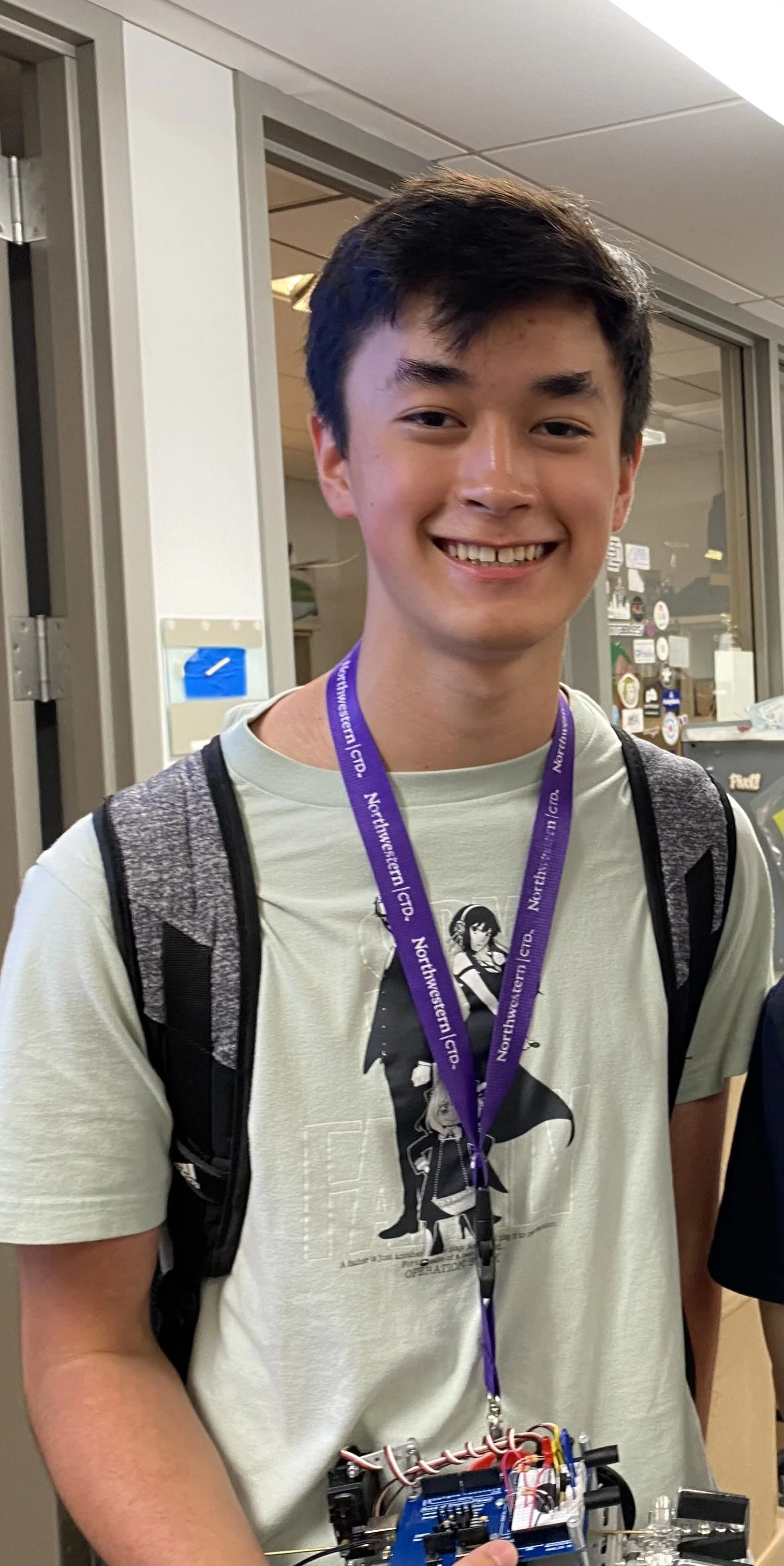 A young man with short dark hair, wearing a gray t-shirt with an anime graphic, a purple lanyard, and a backpack, smiling and holding a small electronic robot.