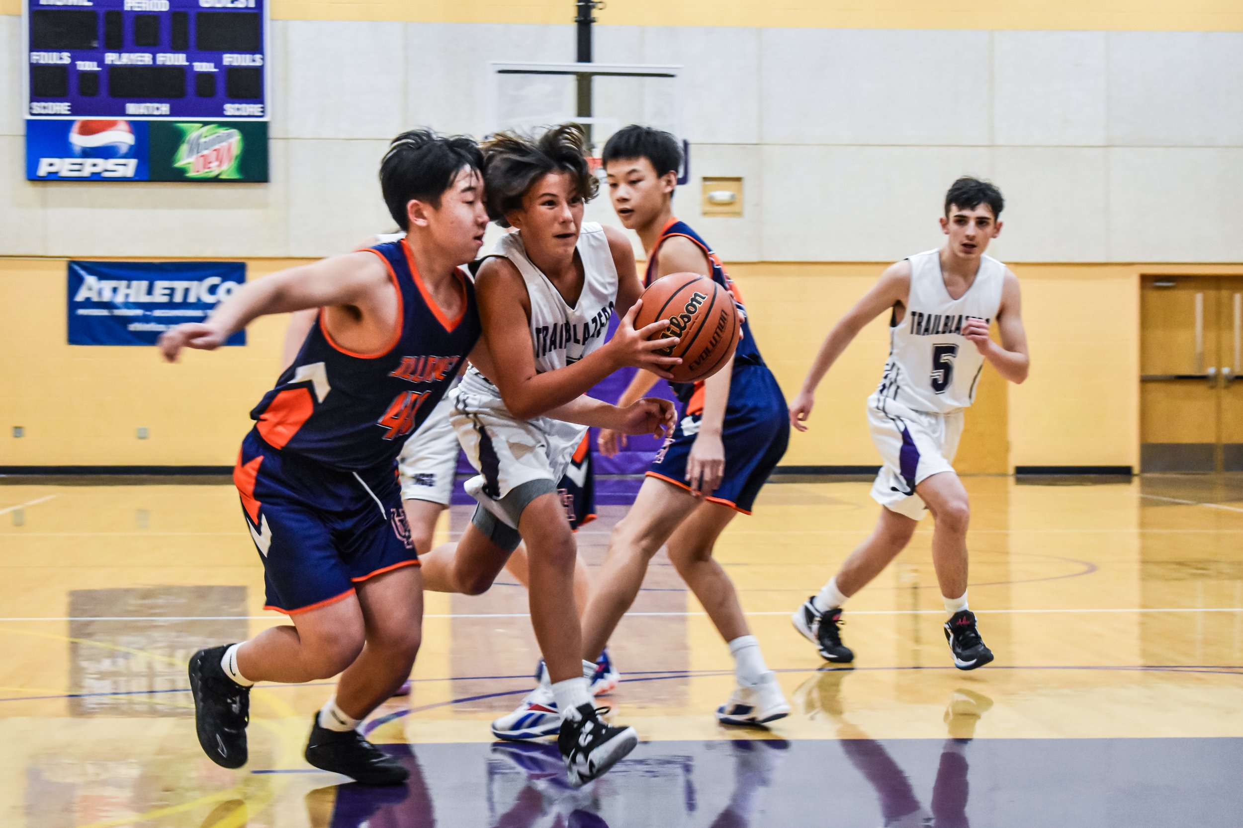 Youth basketball game with three players in white uniforms and two in blue and orange uniforms competing for the ball on a gym court.
