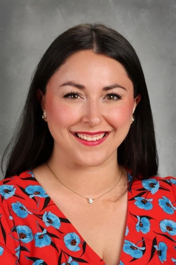 Portrait of a smiling woman with long black hair, wearing a red floral top with blue flowers, earrings, and a delicate necklace against a neutral background.