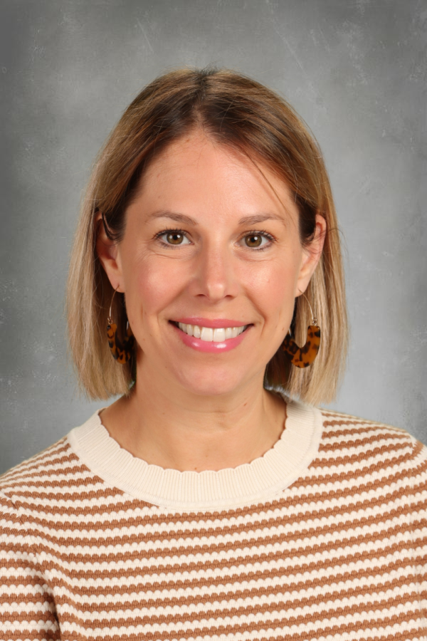 A woman with shoulder-length blonde hair, wearing a cream and brown striped sweater, and tortoiseshell earrings, smiling against a gray background.